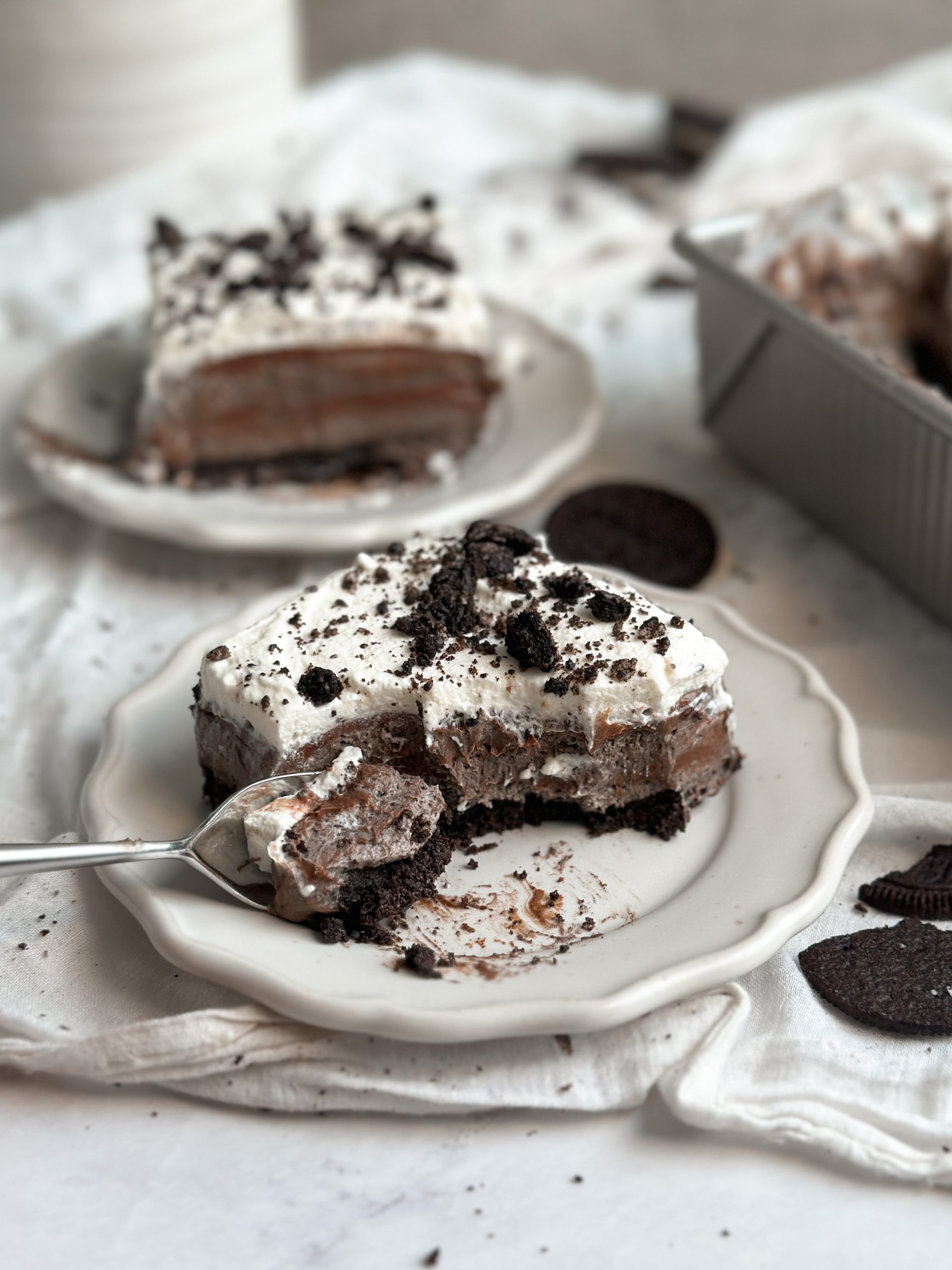 a slice of no bake cookies and cream mousse cake on a small plate with a crust, layers of mousse and ganache, and a spoon is taking out a bite from the cake. another plate in background