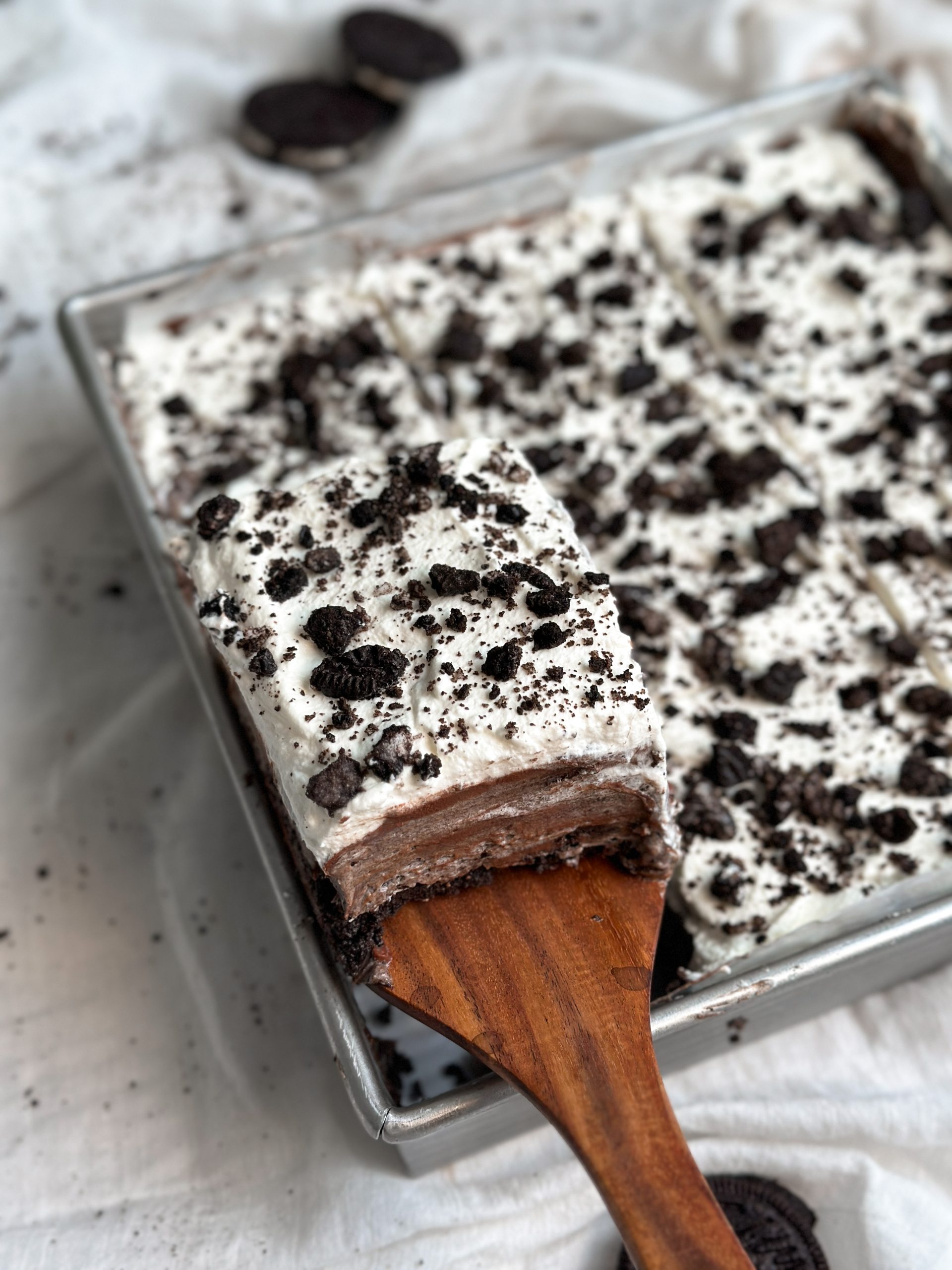 a baking dish with a no bake cookies & cream (oreo) mousse cake, a spatula is pulling out one slice revealing layers of mousse and ganache