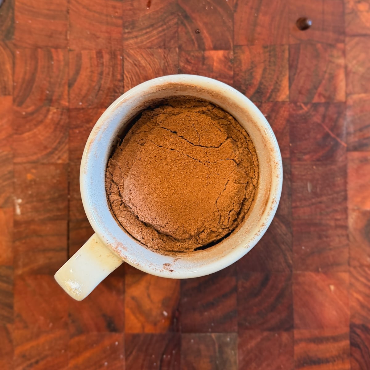 A white mug filled with flour and cocoa powder sits on a wooden cutting board