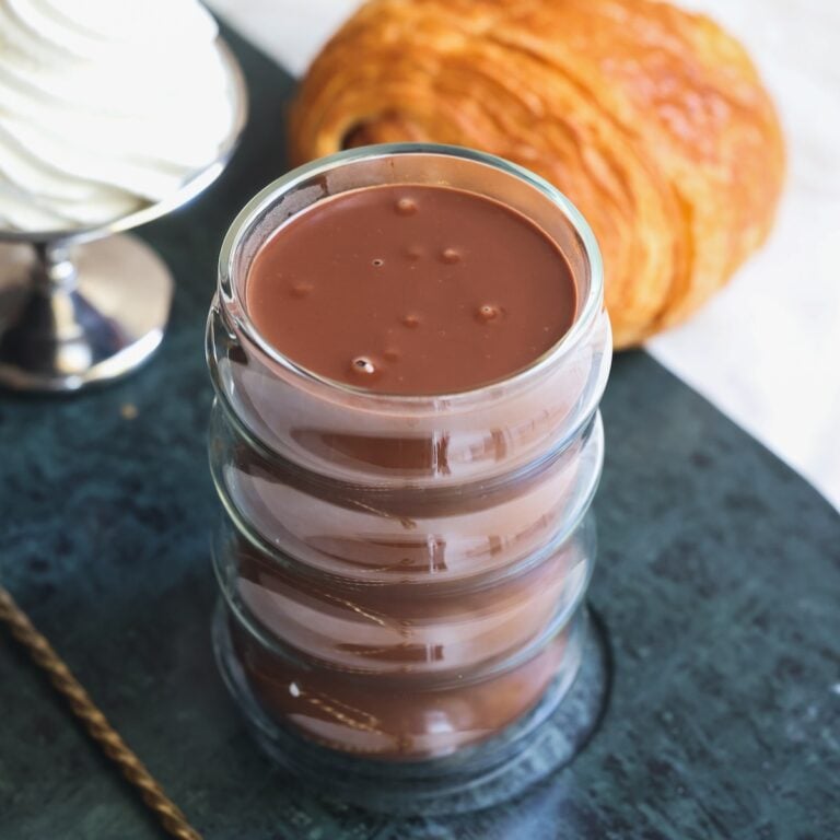 A double-walled glass filled with french hot chocolate, sits on a dark surface, with a whipped cream dessert and a pastry, likely a pain au chocolate, in the background. The hot chocolate appears thick and creamy.