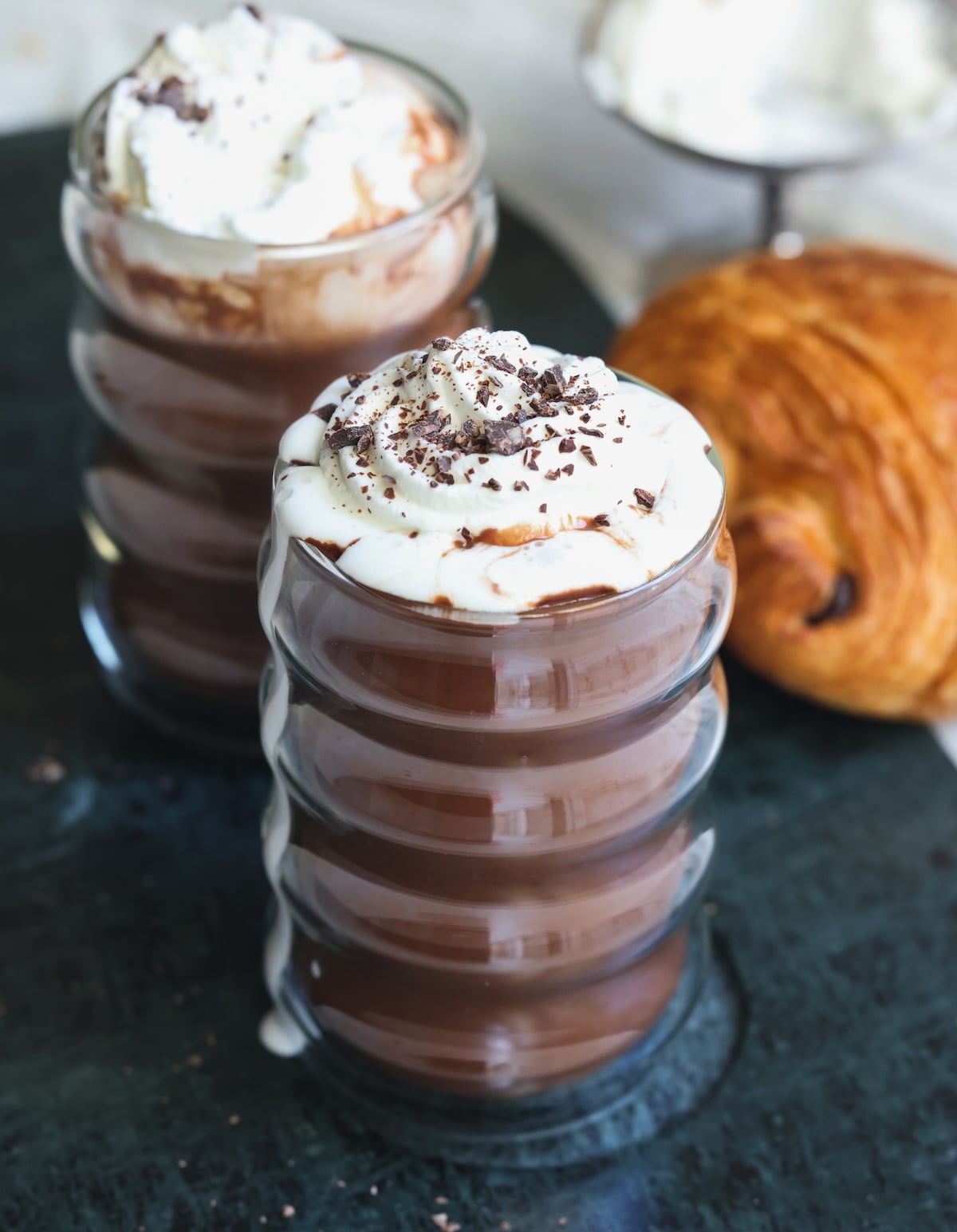 Two glass cups of french hot chocolate topped with whipped cream and chocolate shavings sit on a dark surface, with a croissant and more whipped cream in the background.