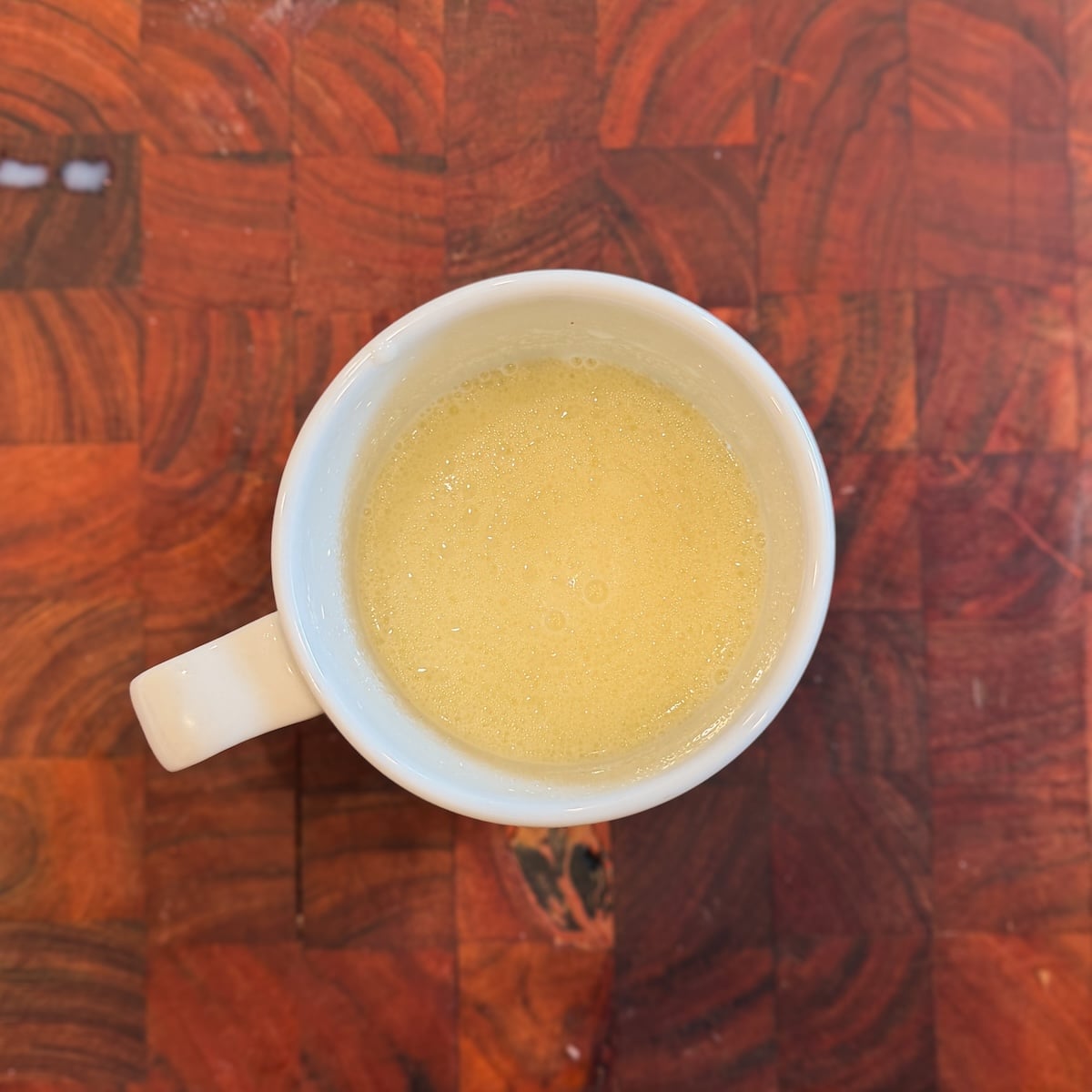 A white mug is filled with a light yellow frothy mixture of liquids and sugar, and sits on a wooden surface with a checkered pattern.