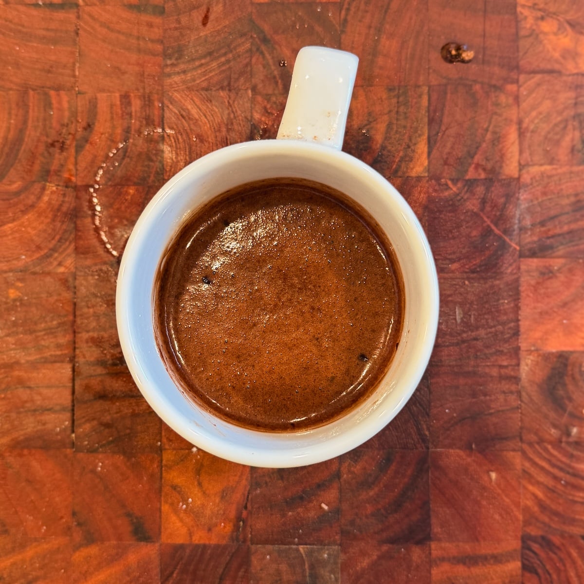 A white cup filled with chocolate cake batter sits on a wooden surface, viewed from above.
