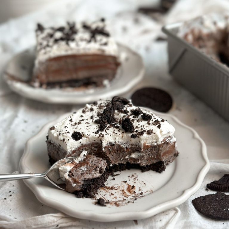 a slice of no bake cookies and cream mousse cake on a small plate with a crust, layers of mousse and ganache, and a spoon is taking out a bite from the cake.