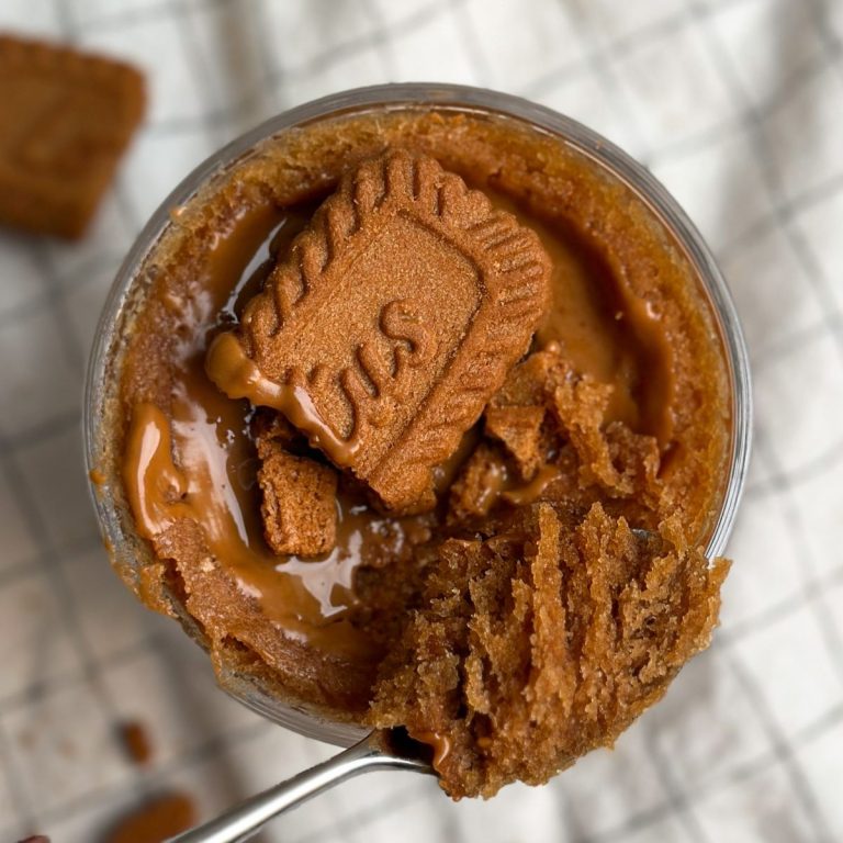 Overhead picture of a Biscoff mug cake in a glass mug, covered in biscoff cookie butter and crushed cookies. spoon taking out a bite revealing moist texture inside