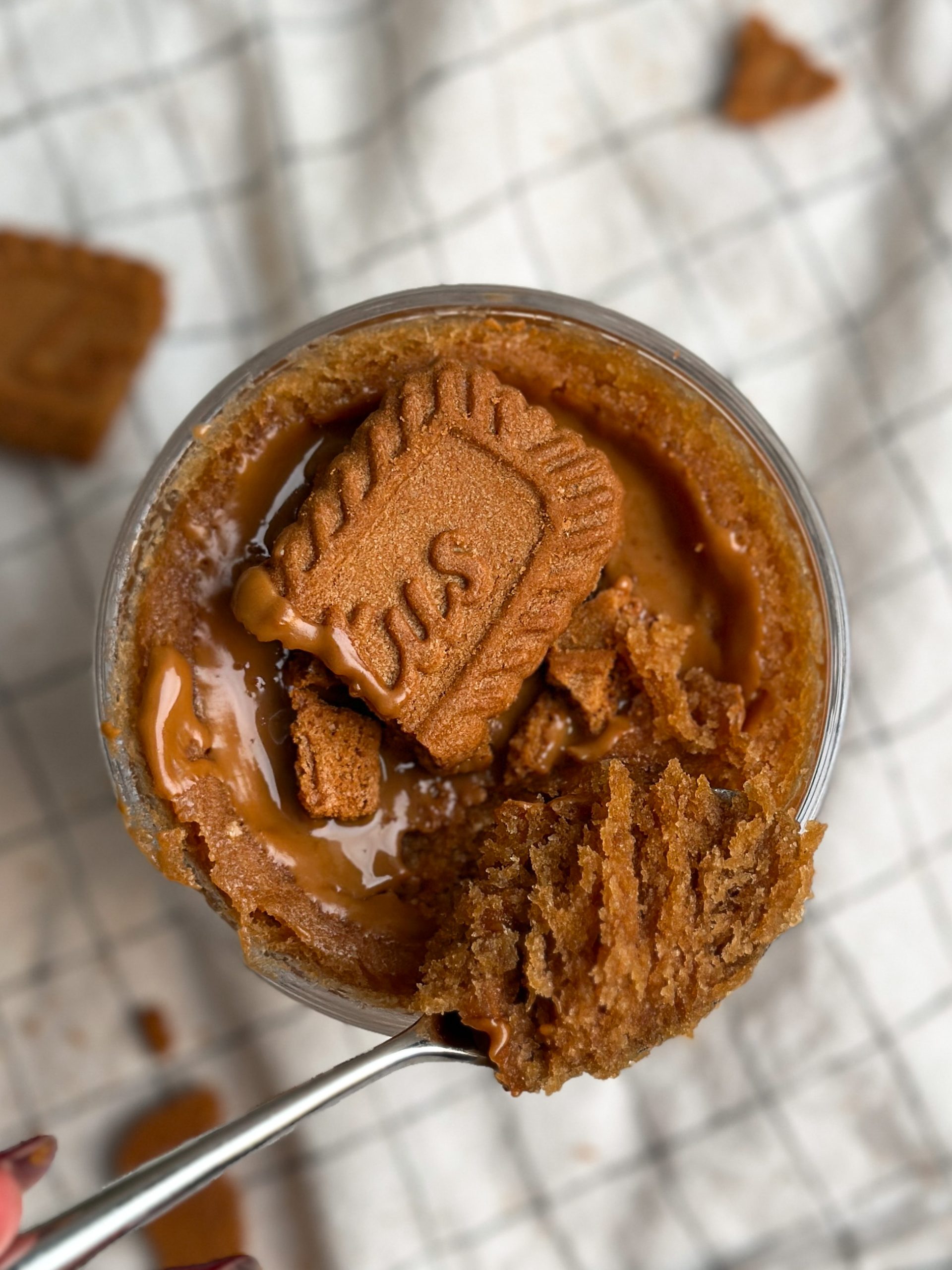 Overhead picture of a Biscoff mug cake in a glass mug, covered in biscoff cookie butter and crushed cookies. spoon taking out a bite revealing moist texture inside
