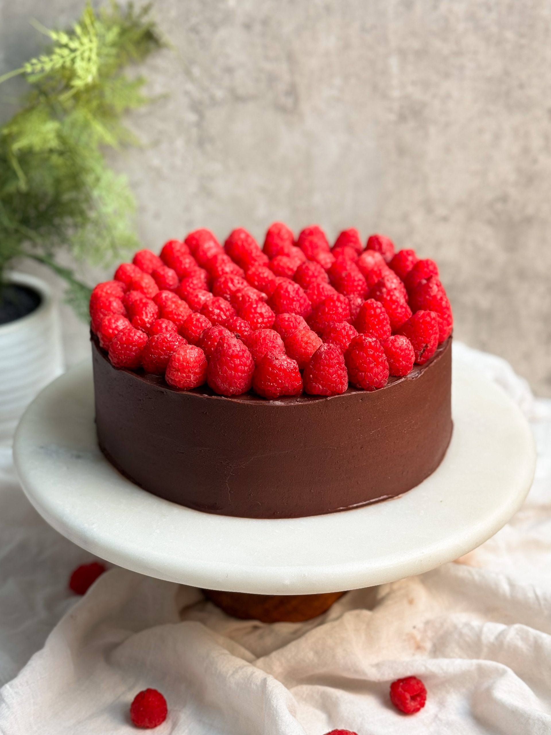 A chocolate cake covered in ganache and decorated with fresh raspberries on a white serving board