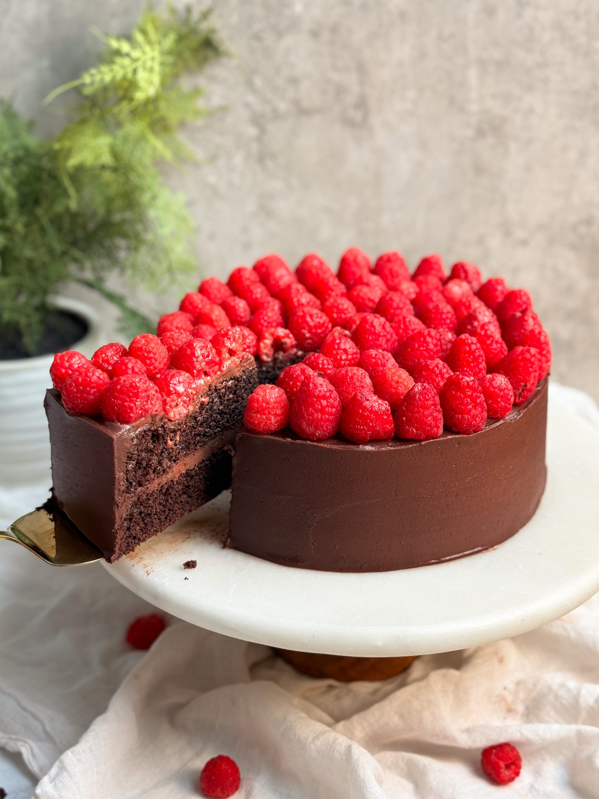 Chocolate raspberry fudge cake on a marble serving board with a spatula pulling out a slice revealing moist chocolatey interior, chocolate ganache, and layer of raspberries on top