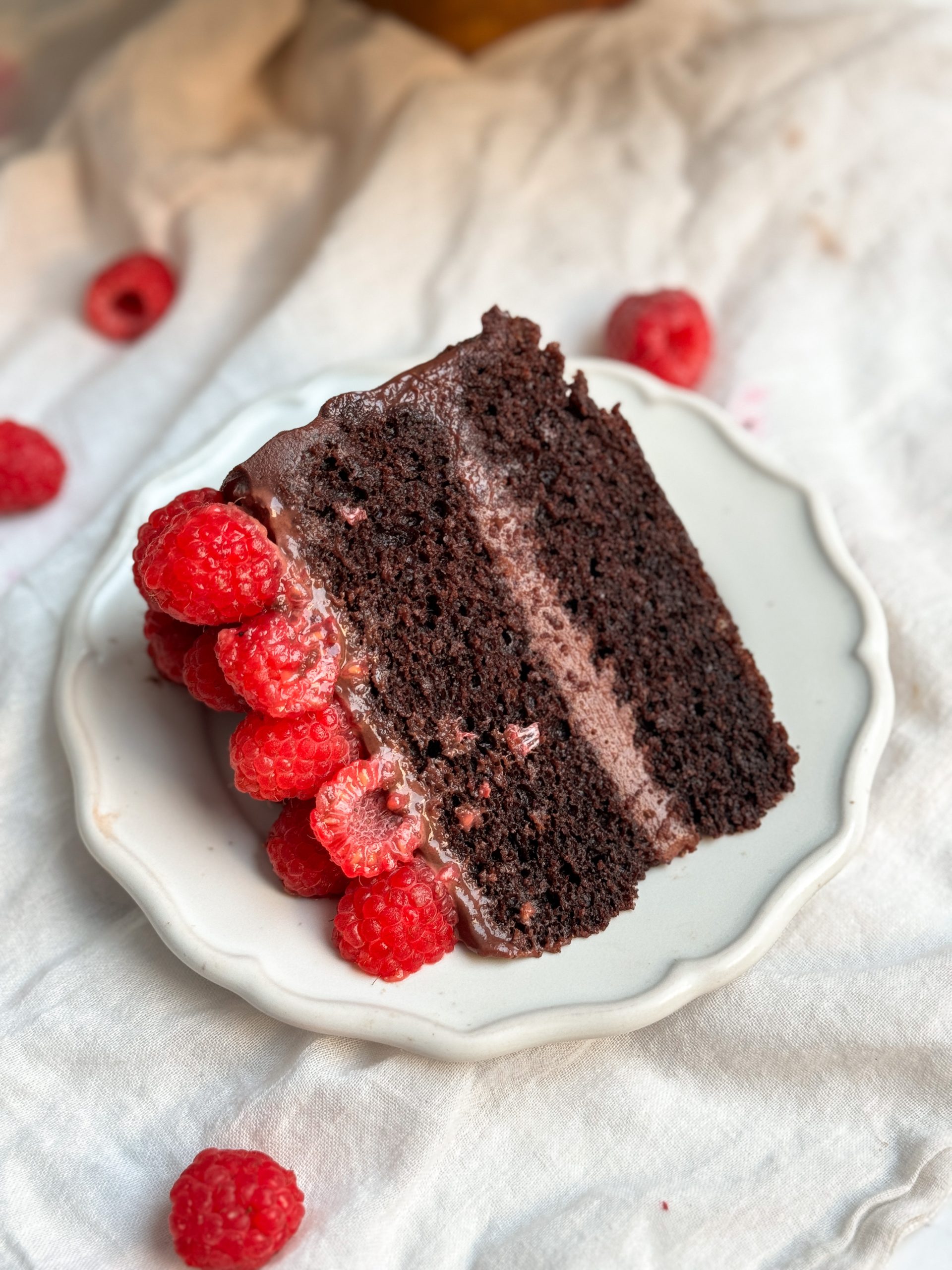 A slice of chocolate raspberry fudge cake on a small plate. cake has 2 layers of moist chocolate cake, ganache in between and on top, and is decorated with raspberries