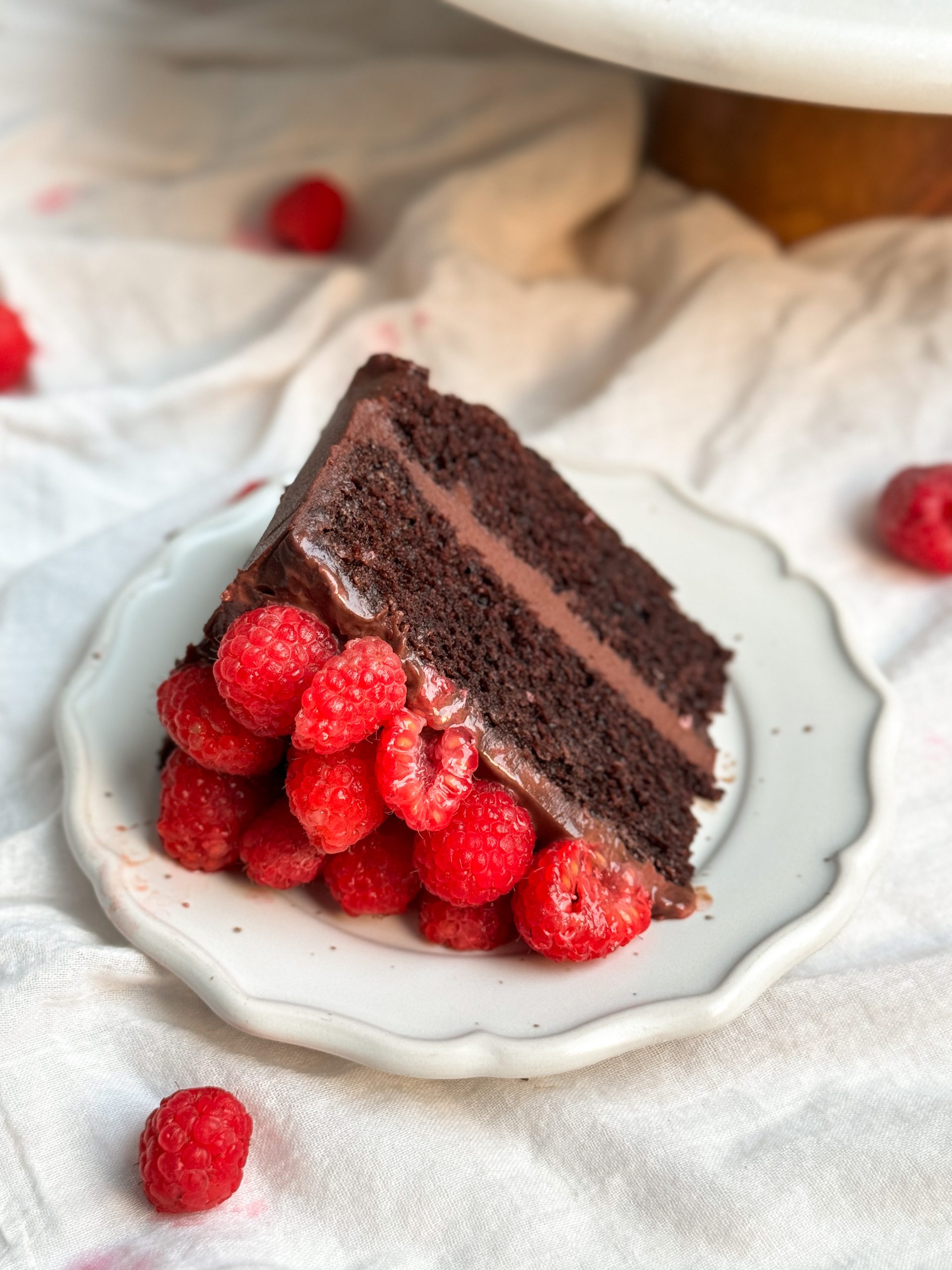 A slice of chocolate raspberry fudge cake on a small plate. cake has 2 layers of moist chocolate cake, ganache in between and on top, and is decorated with raspberries. picture from an angle