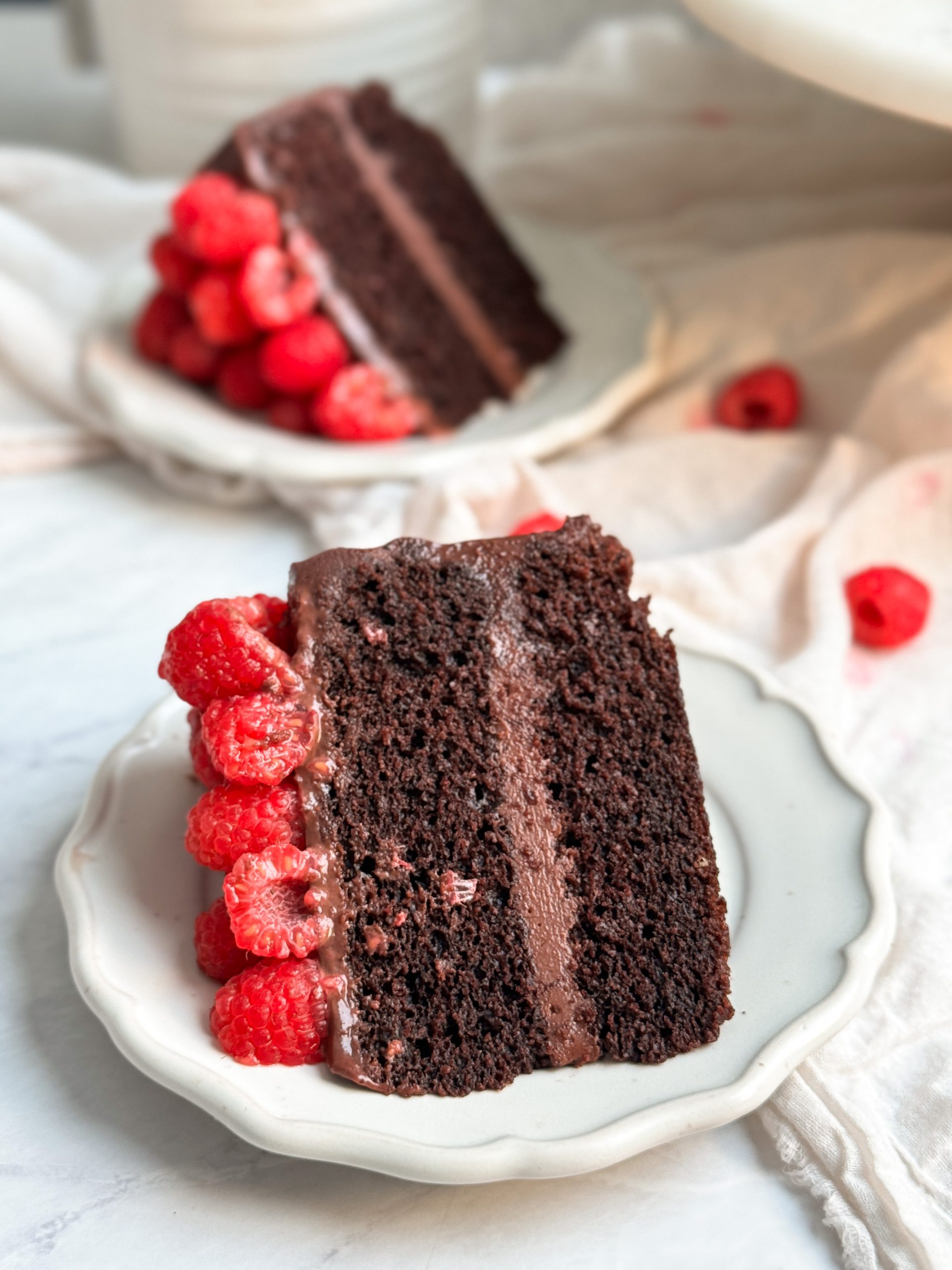 A slice of chocolate raspberry fudge cake on a small plate. cake has 2 layers of moist chocolate cake, ganache in between and on top, and is decorated with raspberries. Another slice seen in the background