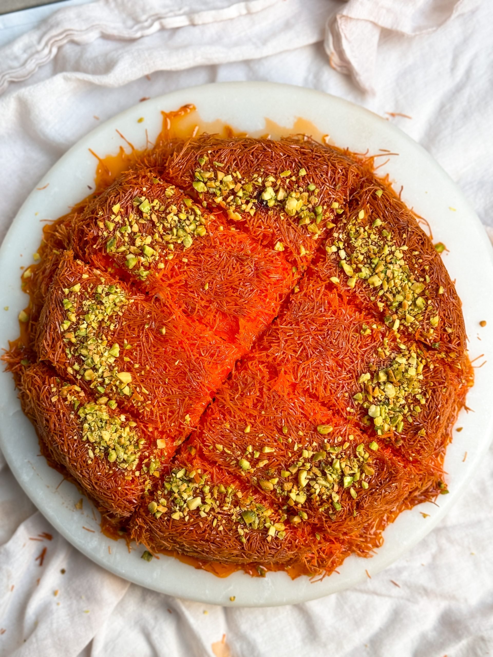 knafeh on a white marble serving board. knafeh has a crispy orange exterior, is soaked in syrup, decorated with chopped pistachios and cut into slices