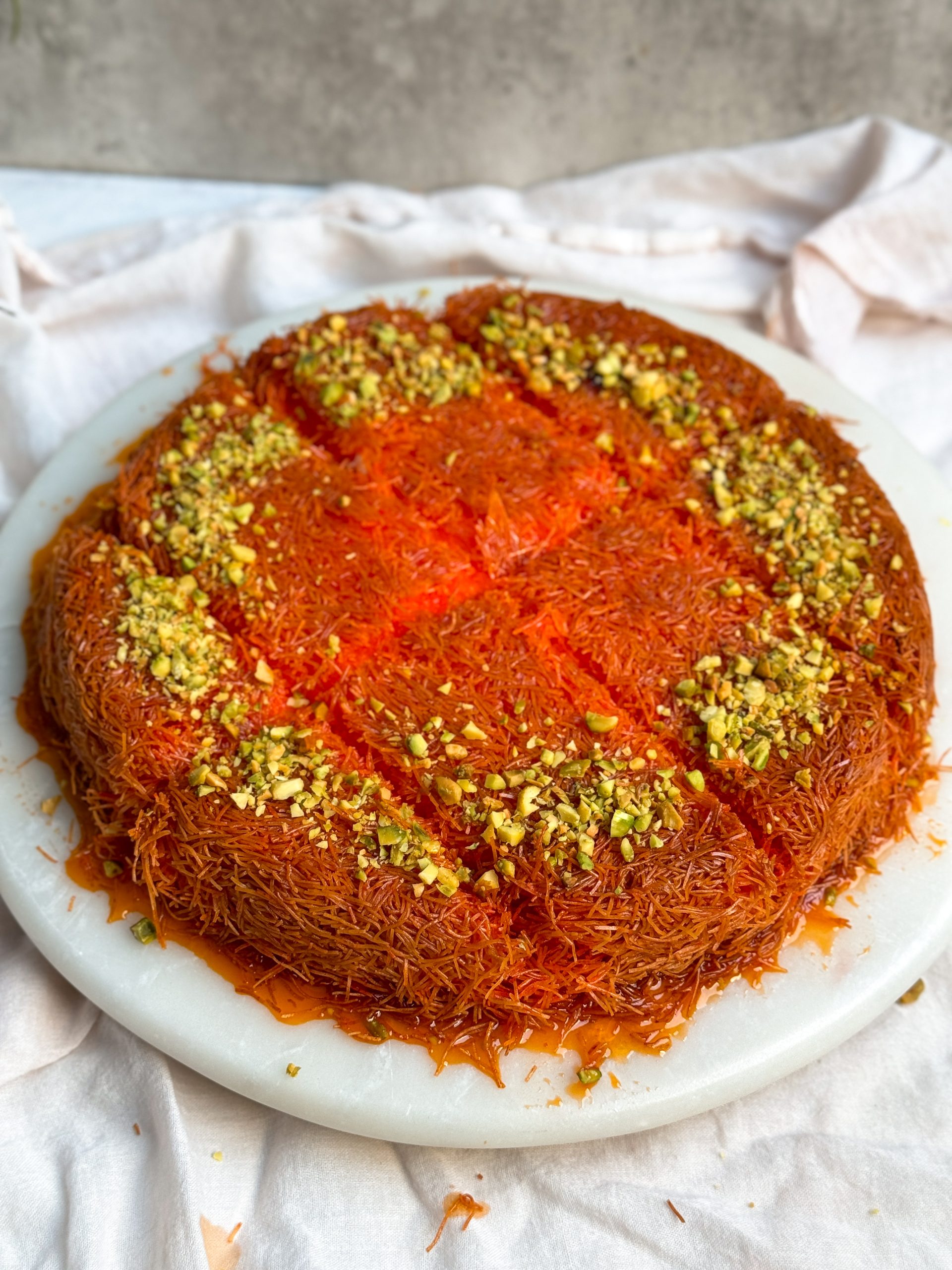 knafeh on a white marble serving board. knafeh has a crispy orange exterior, is soaked in syrup, decorated with chopped pistachios and cut into slices. picture at an angle