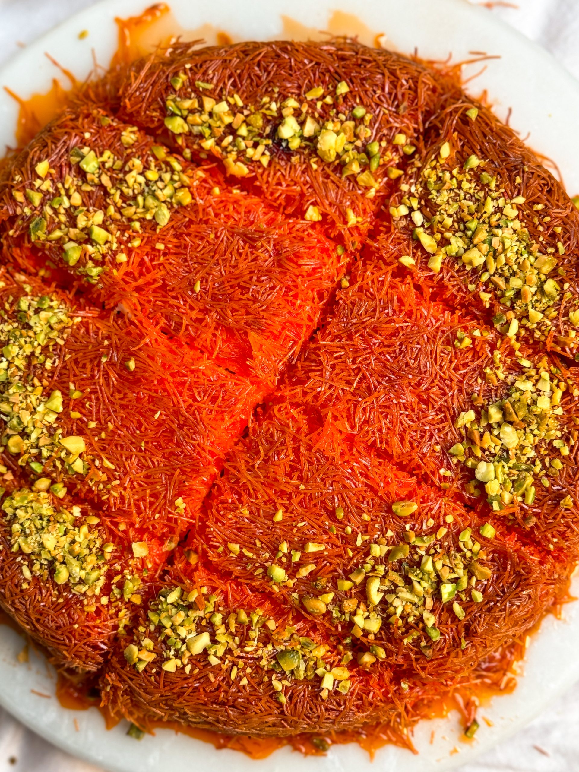 knafeh on a white marble serving board. knafeh has a crispy orange exterior, is soaked in syrup, decorated with chopped pistachios and cut into slices; close up picture