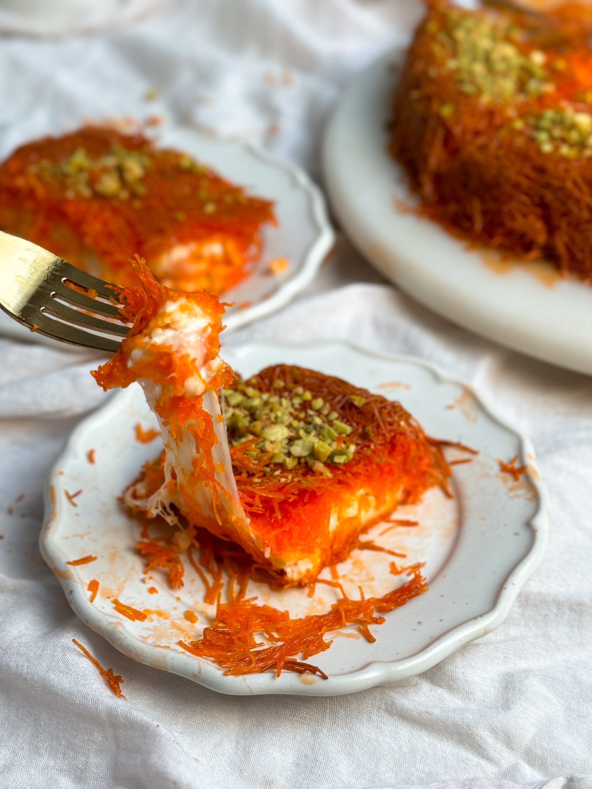 slice of knafeh with crispy pastry and melted cheese inside on a small plate, another plate in the background. fork is pulling out a bite with a string of cheese (cheese pull)