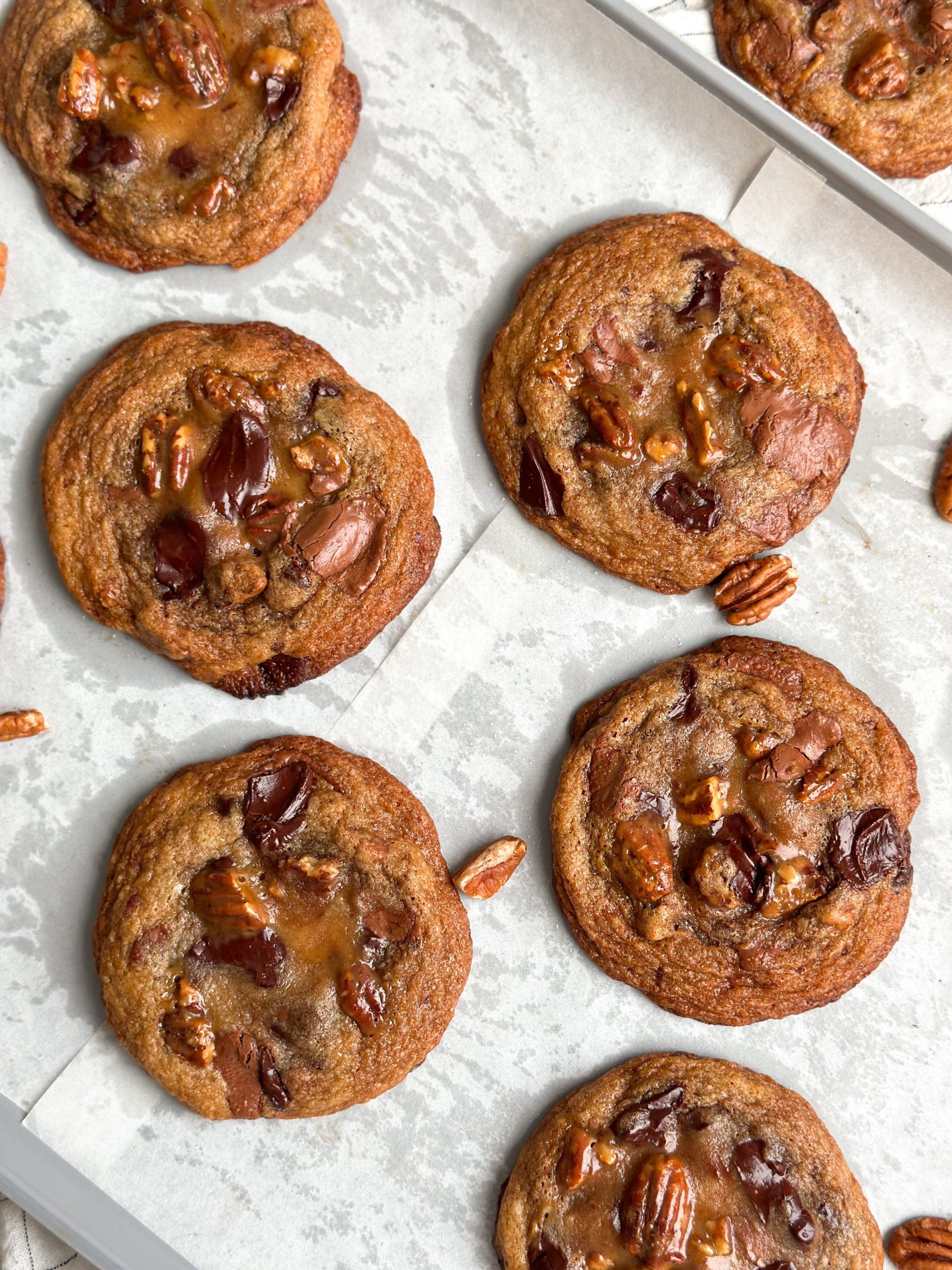 6 salted caramel pecan chocolate chip cookies placed side by side with some pecans scattered. cookies are a deep golden color and look crispy and chewy with puddles of chocolate