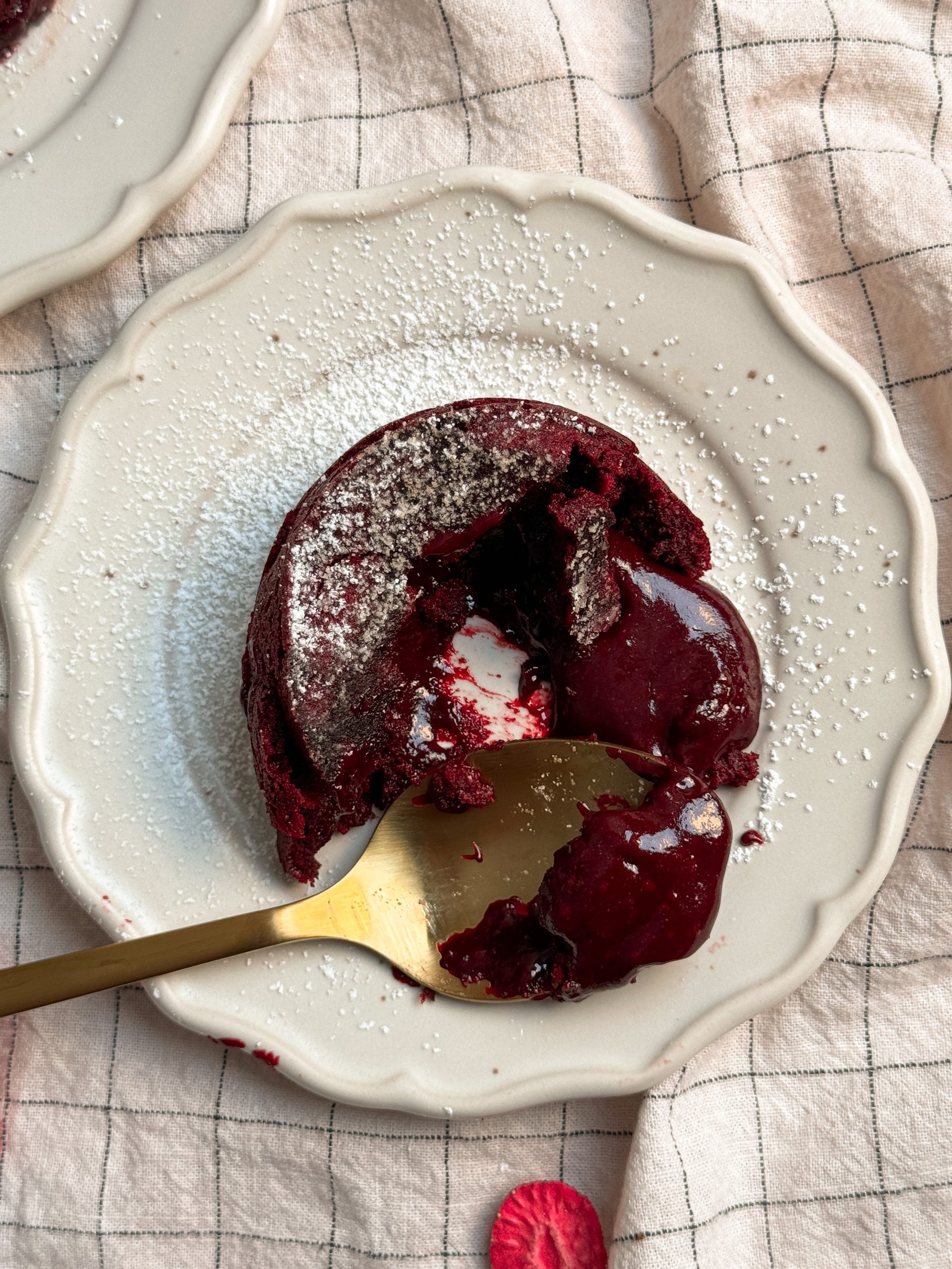red velvet molten lava cake on a small plate. cake is half eaten and the molten center is oozing out, and a spoon is taking a bite out of the cake