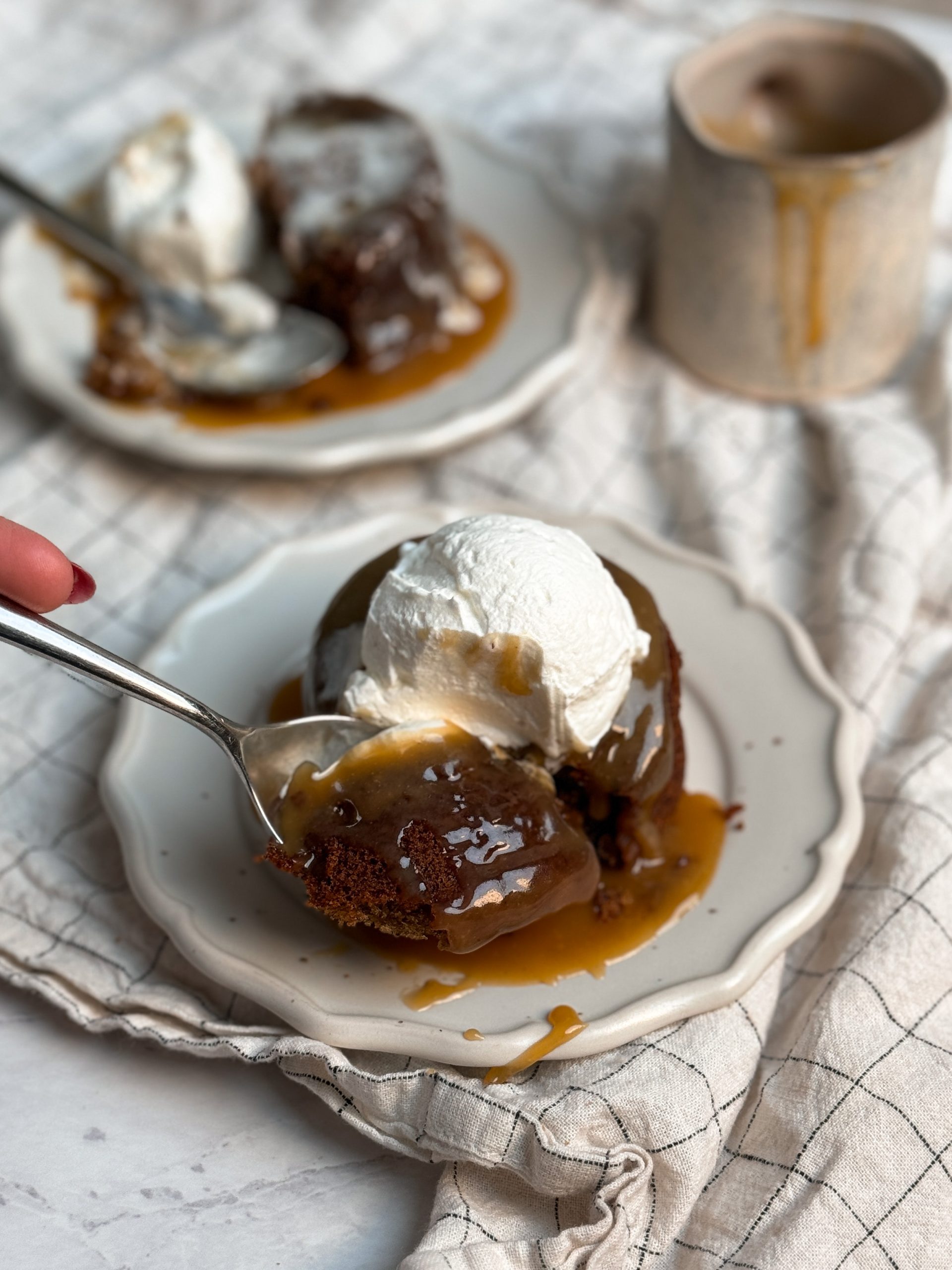 Small dessert place with a single serve sticky toffee pudding cake with toffee sauce and a scoop of cream on it. spoon is taking out a bite revealing soft and moist texture