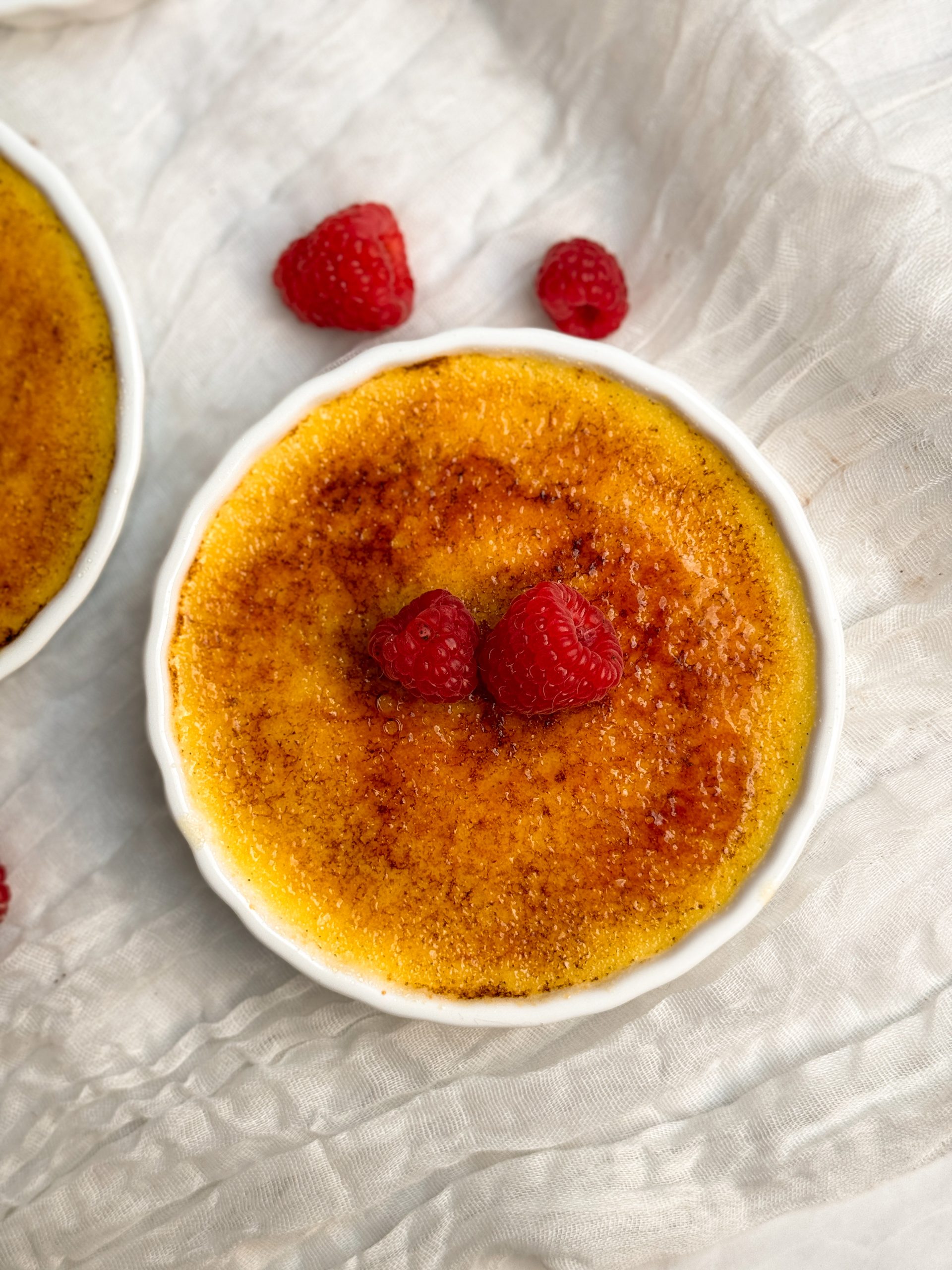 Overhead picture of Creme brulee in a white ramekin with a golden crispy sugar coating, decorated with raspberries