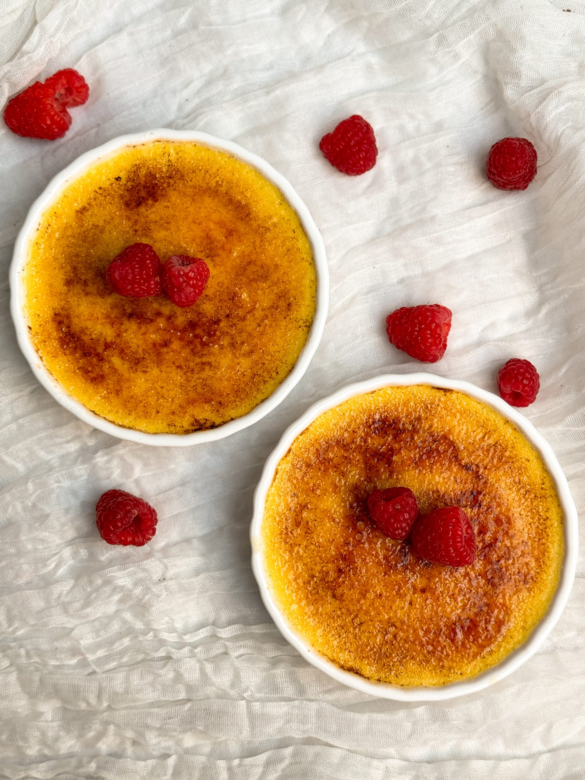 Two creme brulees in a white ramekins with a golden crispy sugar coating, decorated with raspberries