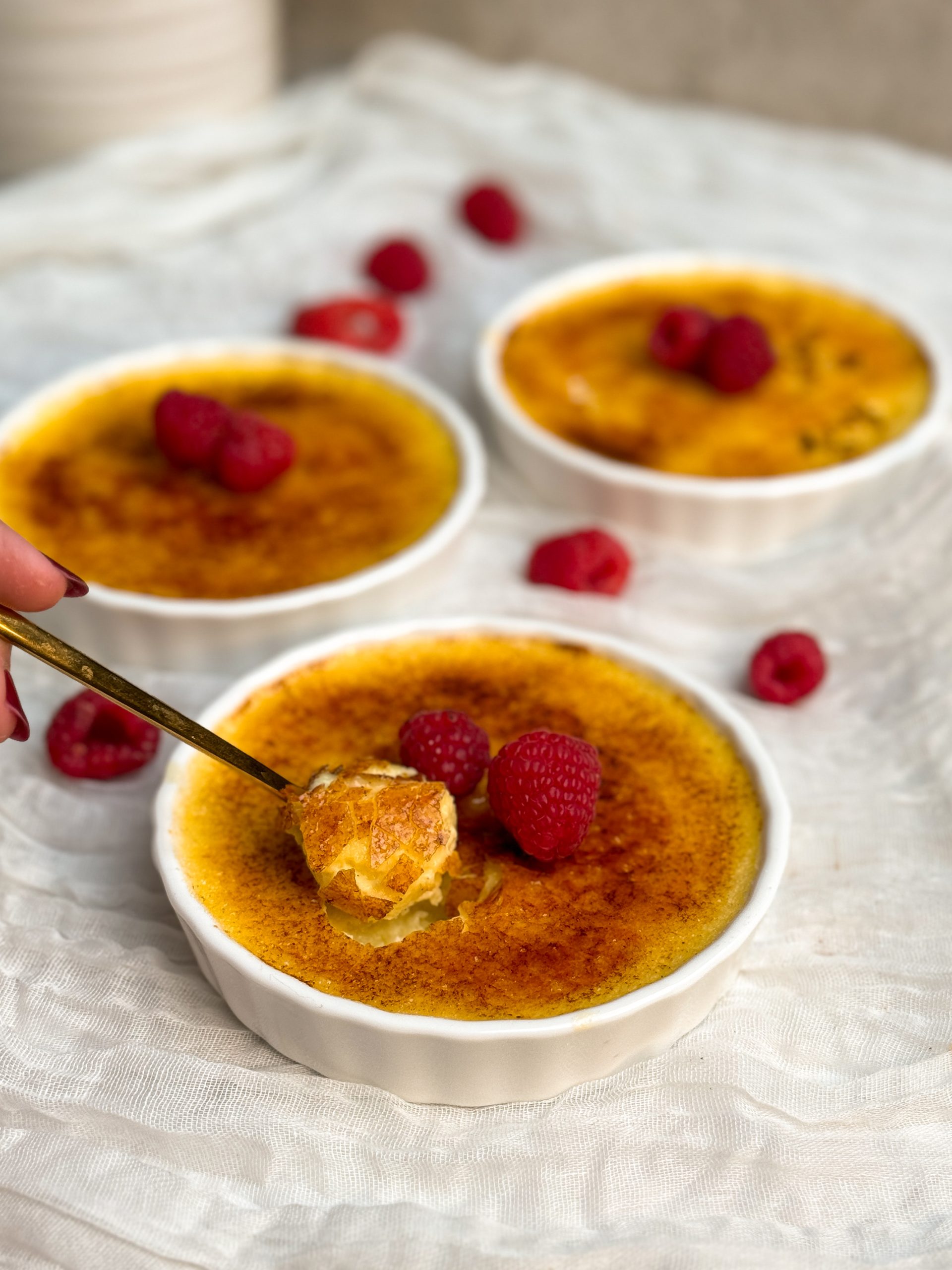 Three creme brulees in a white ramekins with a golden crispy sugar coating, decorated with raspberries. A small golden spoon is taking out a bite from the one in the front showing the thin crispy coating