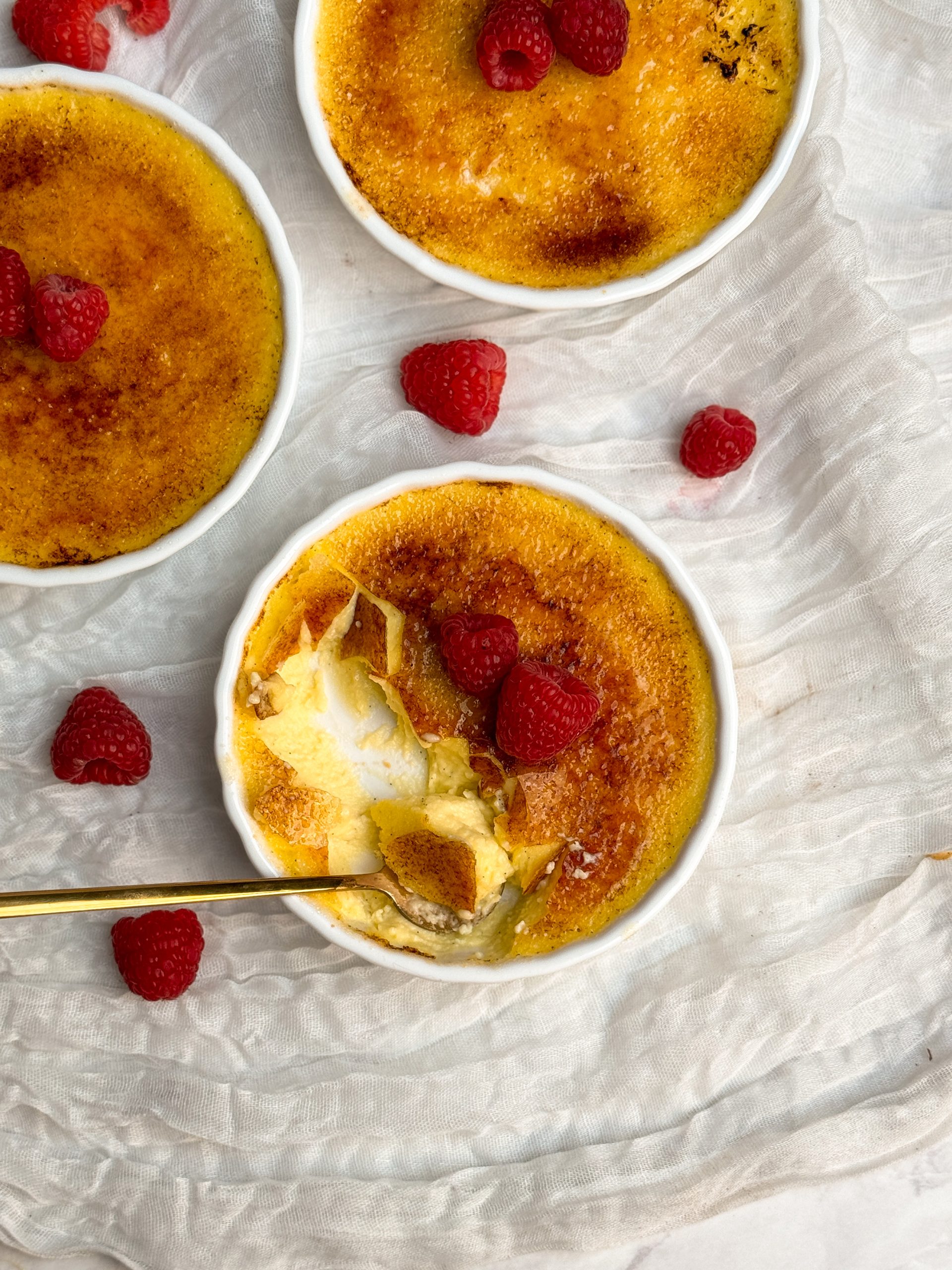 Overhead picture of creme brulee in a white ramekin with a golden crispy sugar coating, deocrated with 2 raspberries. a small golden spoon is taking out a bite showing the thin crispy coating and silky interior. more of the creme brulee is eaten and 2 more are seen on the side