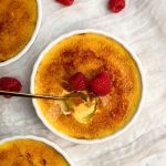 Overhead picture of creme brulee in a white ramekin with a golden crispy sugar coating, deocrated with 2 raspberries. a small golden spoon is taking out a bite showing the thin crispy coating and silky interior