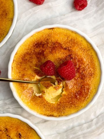 Overhead picture of creme brulee in a white ramekin with a golden crispy sugar coating, deocrated with 2 raspberries. a small golden spoon is taking out a bite showing the thin crispy coating and silky interior