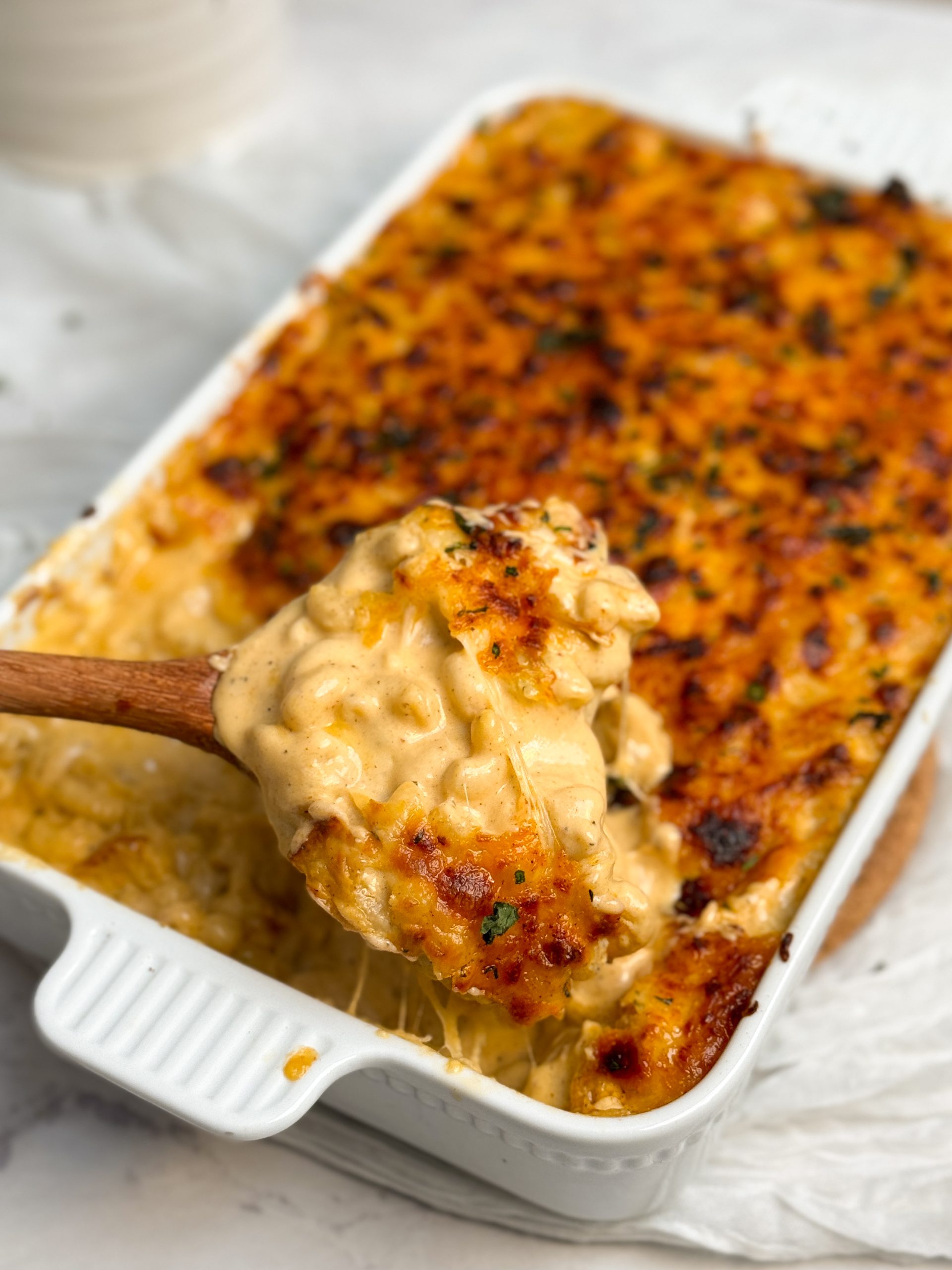 baked mac and cheese in a white casserole dish, mac and cheese is covered by a layer of golden melted cheese. A wooden spoon is scooping out some mac and cheese showing the creamy cheesy texture inside. picture from an angle