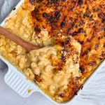 baked mac and cheese in a white casserole dish, picture is close up showing part of it. mac and cheese is covered by a layer of golden melted cheese. A wooden spoon is scooping out some mac and cheese showing the creamy cheesy texture inside