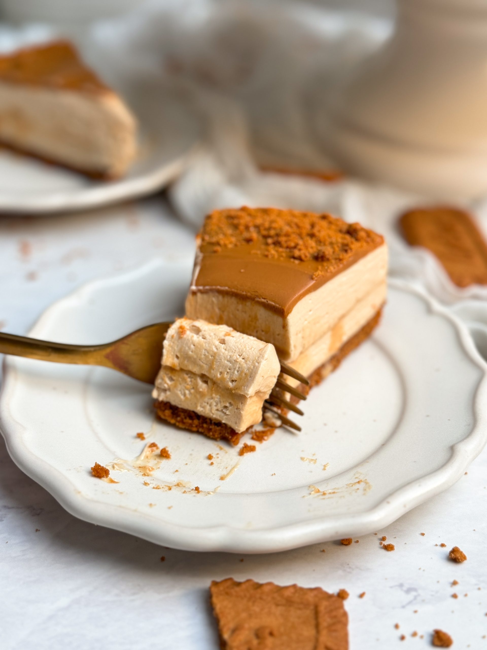 A slice of biscoff salted caramel cheesecake on a small plate with biscoff cookie butter on top. a fork is taking a bite out of the cheesecake. layer of salted caramel in the middle