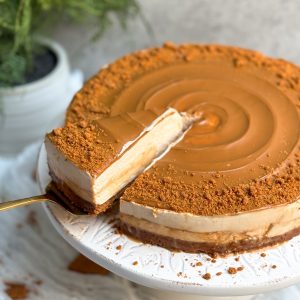 A biscoff salted caramel cheesecake with swirled biscoff cookie butter and crushed cookies on it. cake is on a white cake stand. Some caramel sauce is oozing out from the sides. a spatula is pulling out a slice from the side