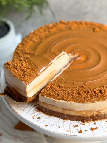 A biscoff salted caramel cheesecake with swirled biscoff cookie butter and crushed cookies on it. cake is on a white cake stand. Some caramel sauce is oozing out from the sides. a spatula is pulling out a slice from the side