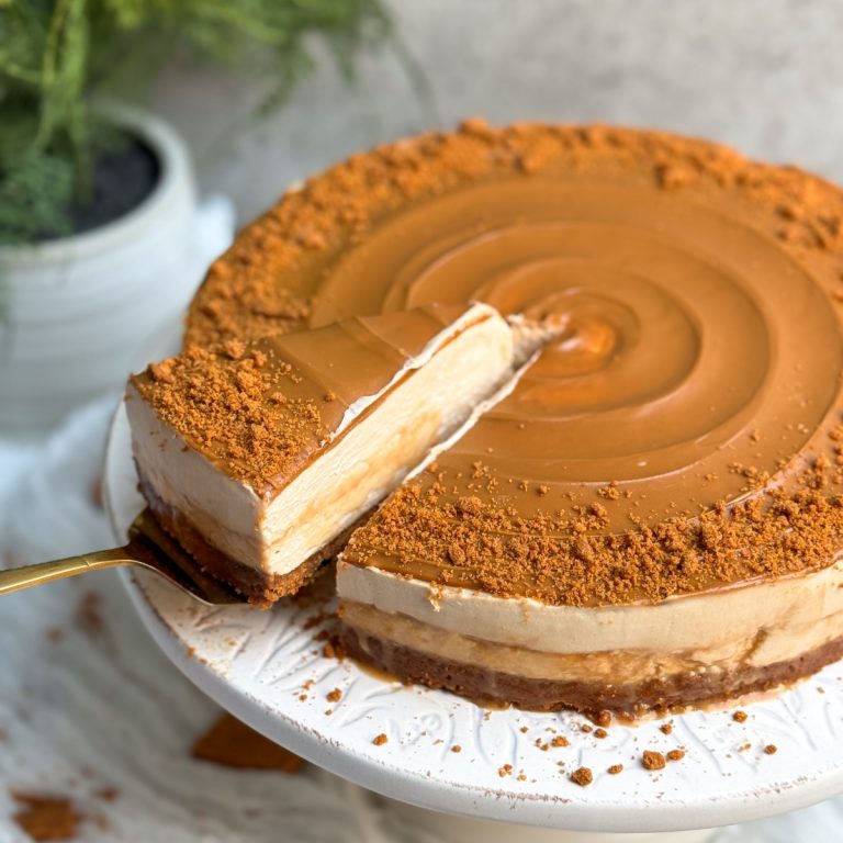 A biscoff salted caramel cheesecake with swirled biscoff cookie butter and crushed cookies on it. cake is on a white cake stand. Some caramel sauce is oozing out from the sides. a spatula is pulling out a slice from the side