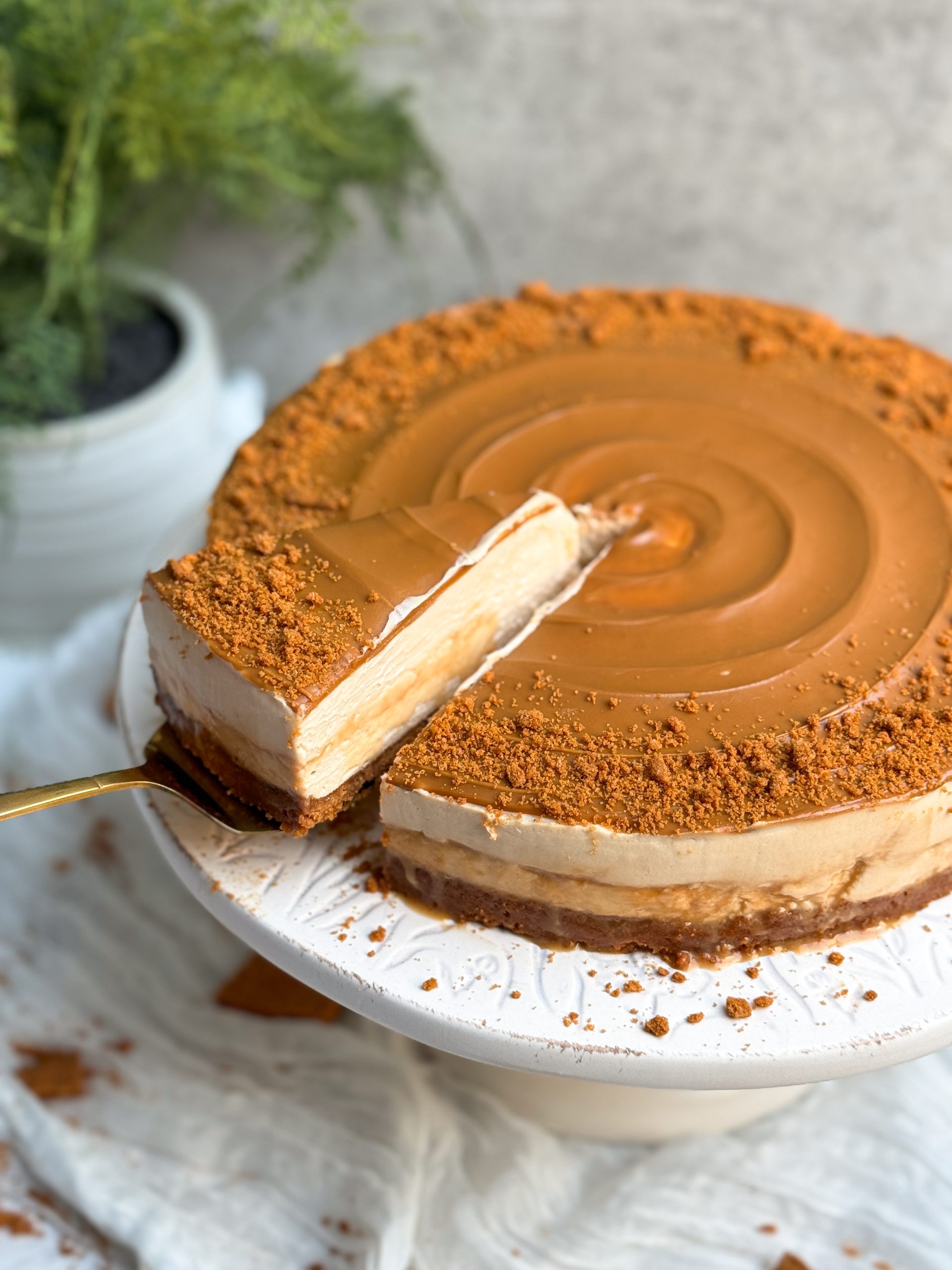 A biscoff salted caramel cheesecake with swirled biscoff cookie butter and crushed cookies on it. cake is on a white cake stand. Some caramel sauce is oozing out from the sides. a spatula is pulling out a slice from the side