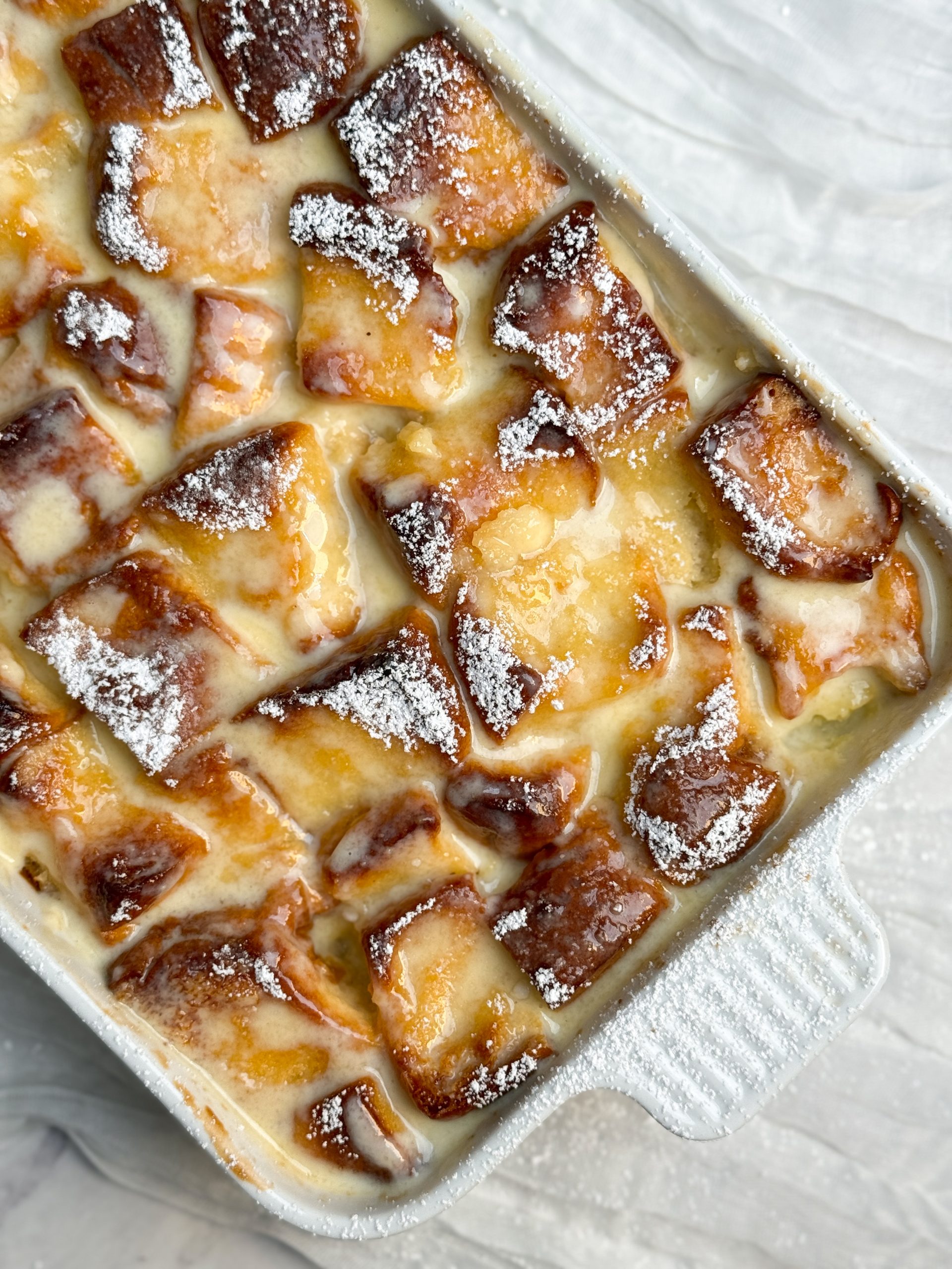 vanilla bread pudding in a white ceramic dish. bread pudding has a lovely golden color and has been soaked in a creme anglaise and dusted with powdered sugar. close up image