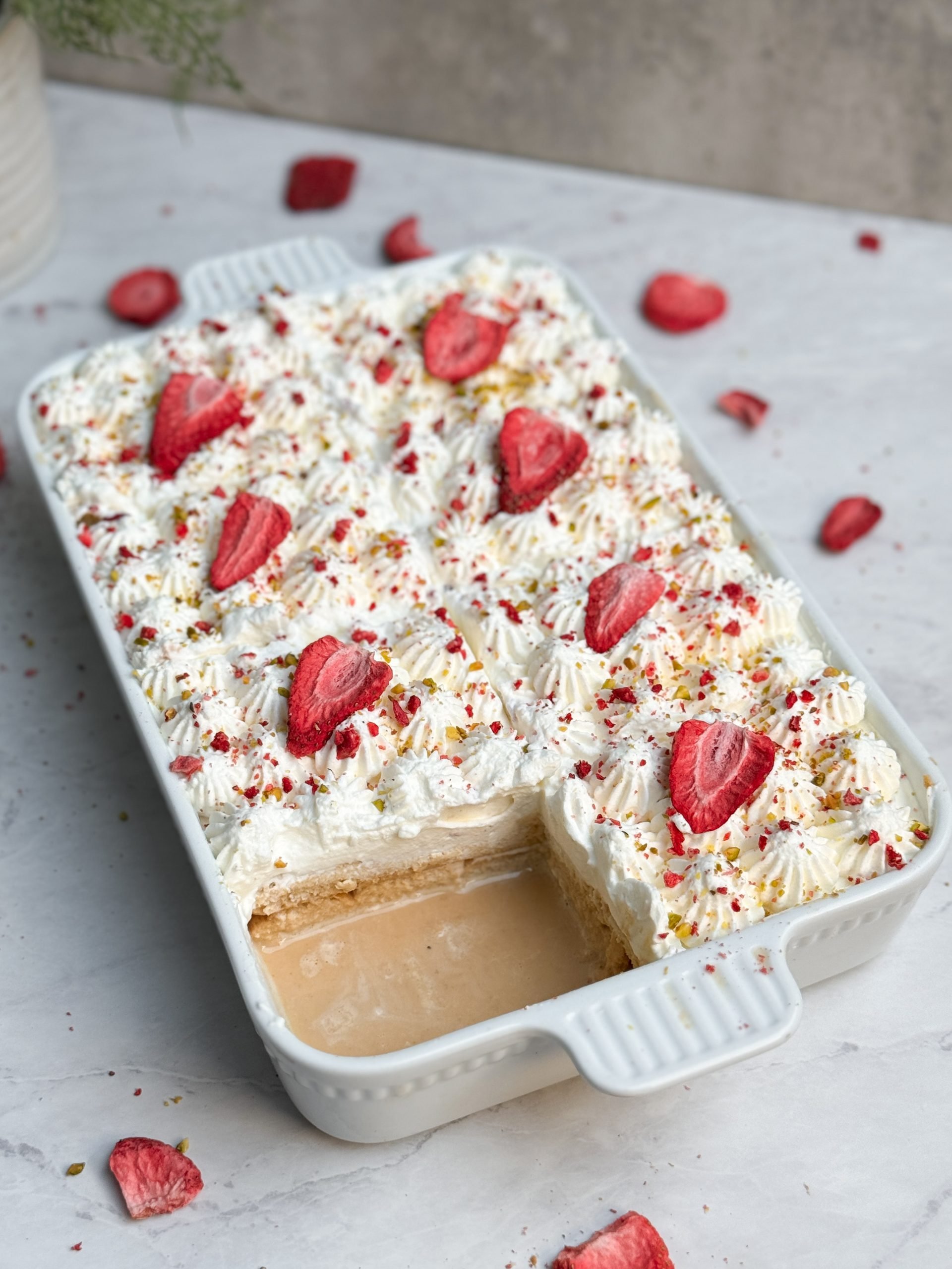 No bake chai tres leches cake in a rectangular ceramic dish decorated with piped whipped cream, freeze dried strawberries and pistachios, one slic eremoved to reveal moist chai soaked interior