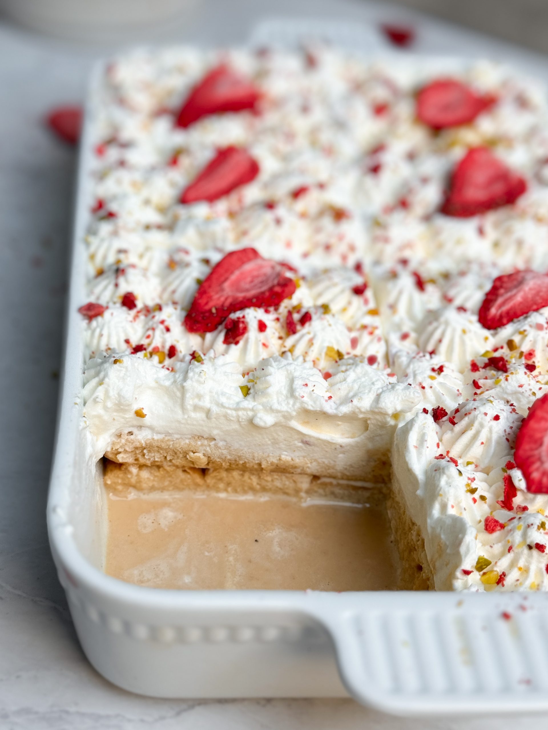 No bake chai tres leches cake in a rectangular ceramic dish decorated with piped whipped cream, freeze dried strawberries and pistachios, one slic eremoved to reveal moist chai soaked interior. close up image