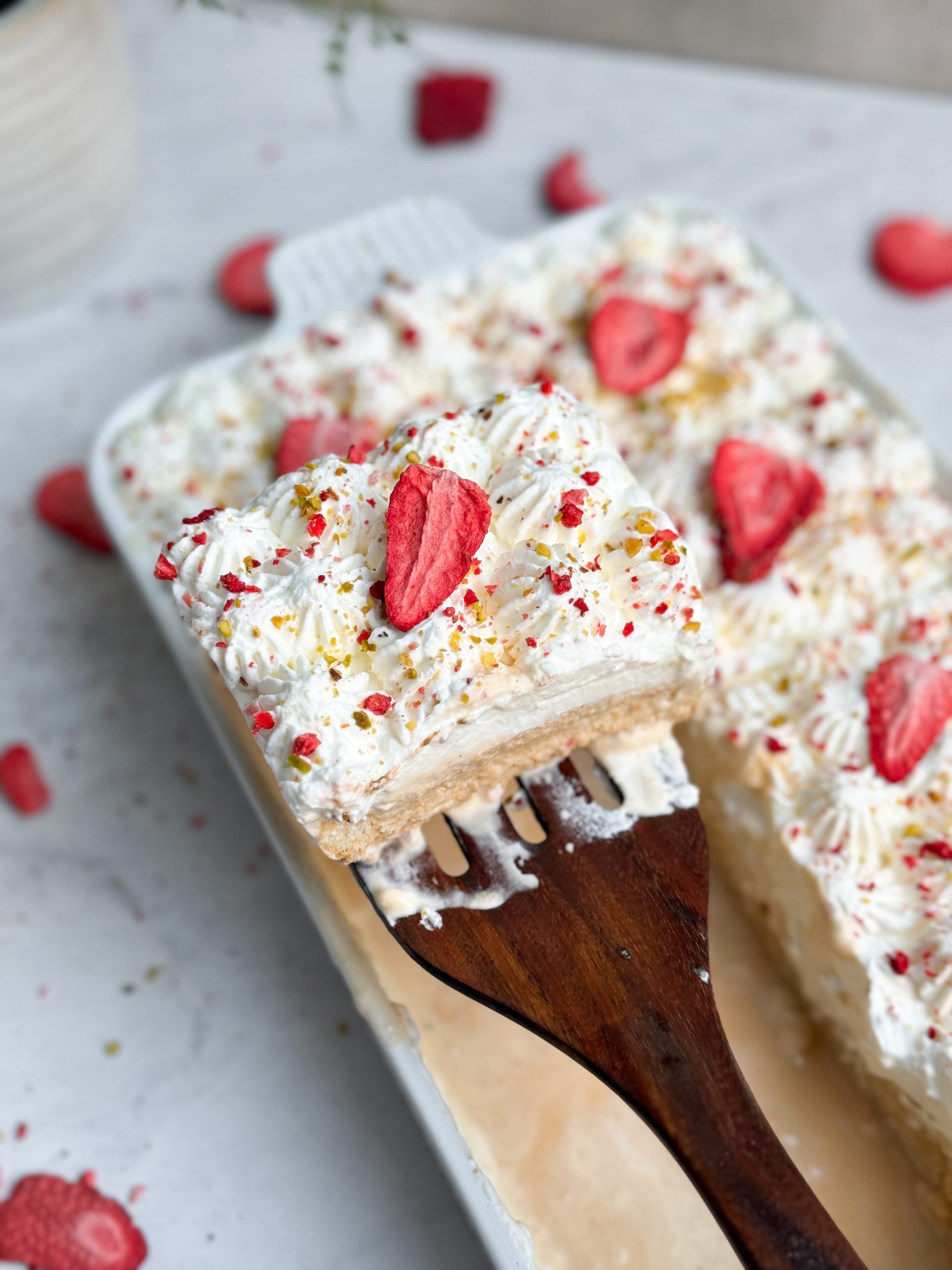 No bake chai tres leches cake in a rectangular ceramic dish decorated with piped whipped cream, freeze dried strawberries and pistachios. The cake is sliced and a spatula is pulling out a slice, picture from the side showing the moist, soaked texture of the cake