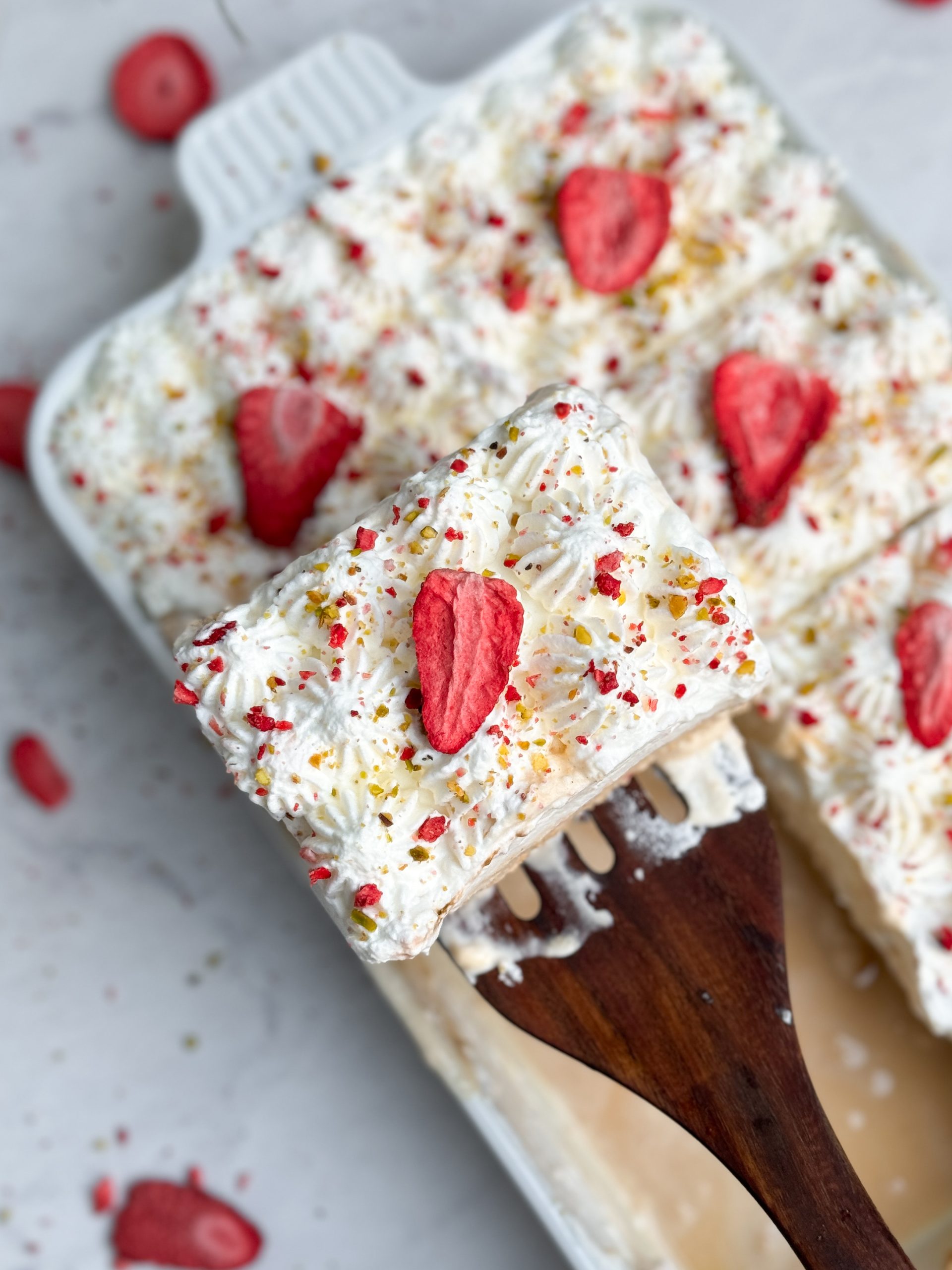 No bake chai tres leches cake in a rectangular ceramic dish decorated with piped whipped cream, freeze dried strawberries and pistachios. The cake is sliced and a spatula is pulling out a slice, picture from the top