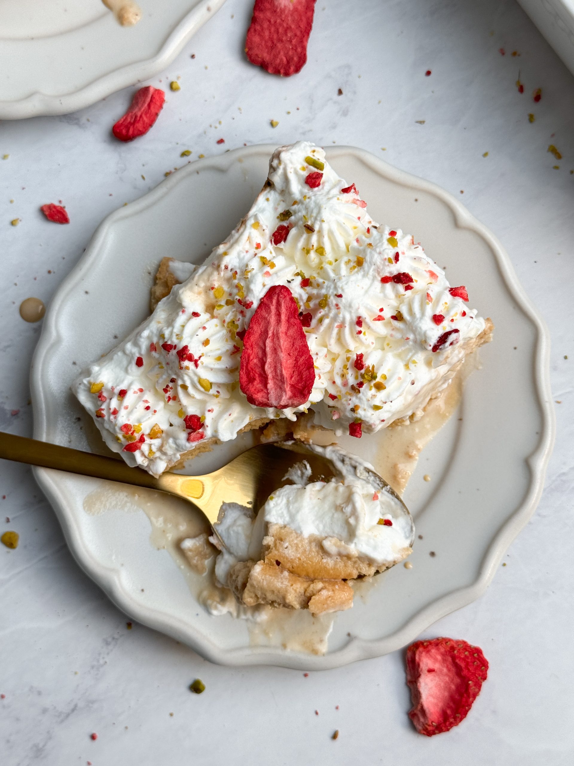 a slice of chai tres leches cake on a small plate decorated with piped whipped cream, freeze dried strawberries and pistachios; spoon is taking out a bite from the plate. picture from the top