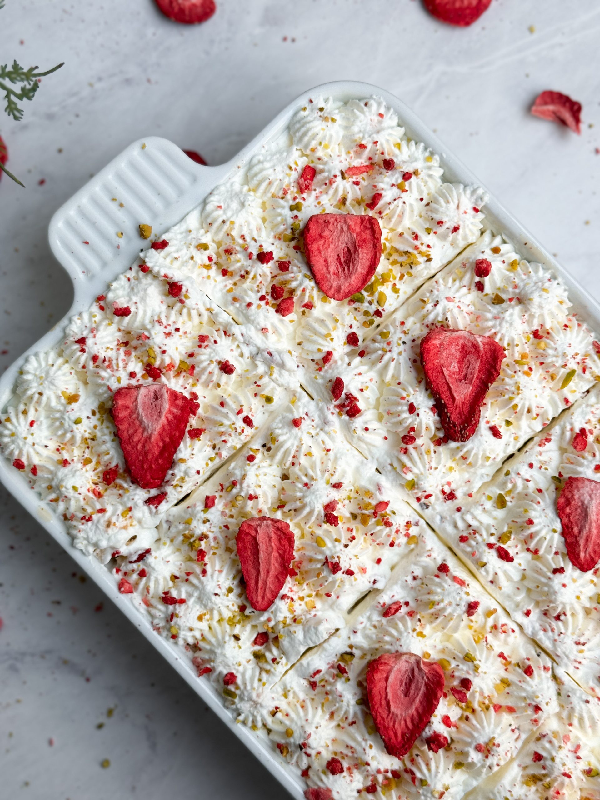No bake chai tres leches cake in a rectangular ceramic dish decorated with piped whipped cream, freeze dried strawberries and pistachios; close up picture