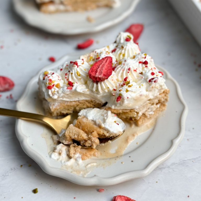 a slice of chai tres leches cake on a small plate decorated with piped whipped cream, freeze dried strawberries and pistachios; spoon is taking out a bite from the plate