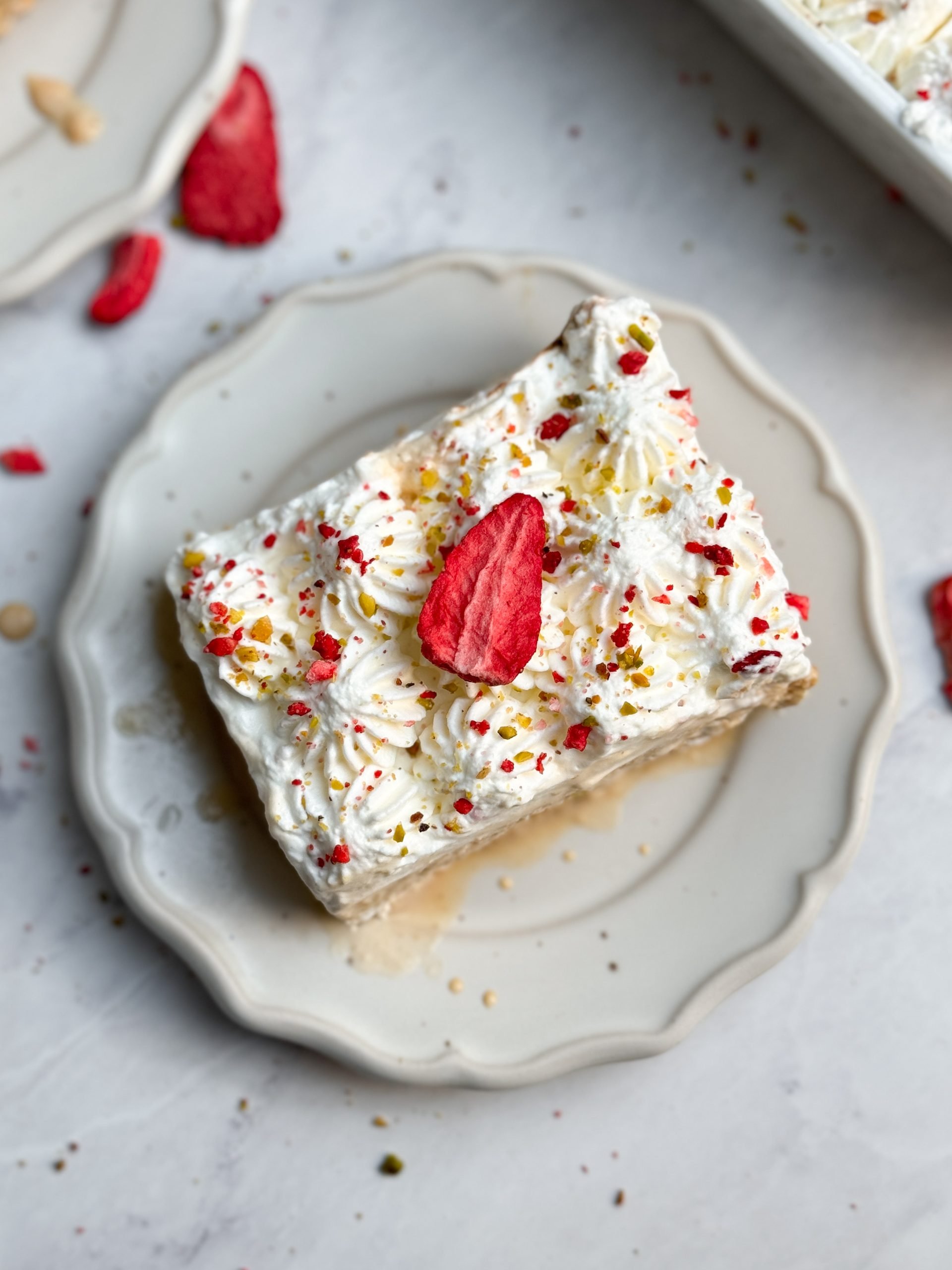 a slice of chai tres leches cake on a small plate decorated with piped whipped cream, freeze dried strawberries and pistachios