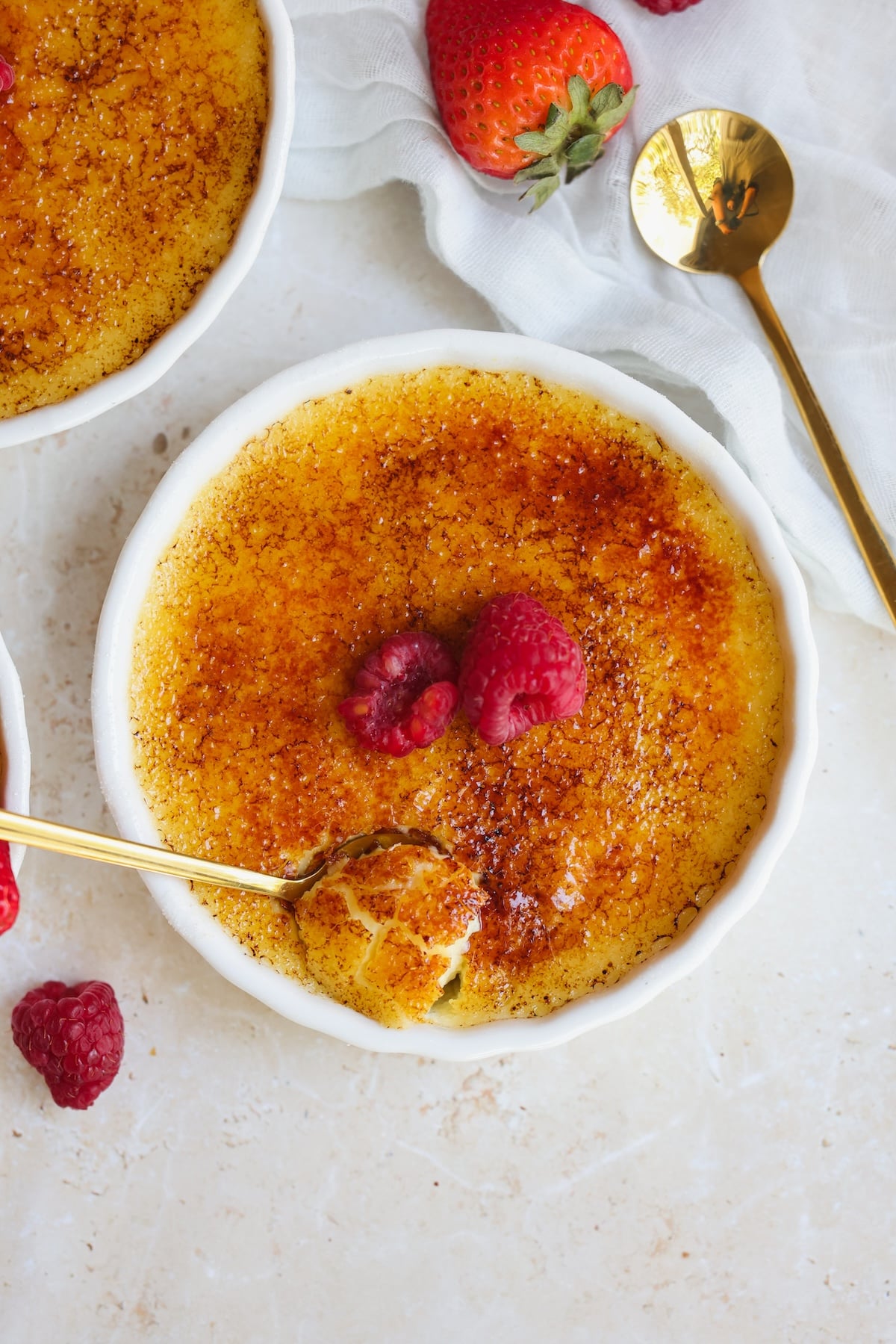 A bowl of crème brûlée with a caramelized sugar top, garnished with raspberries. A gold spoon has cracked the surface, revealing creamy custard inside.