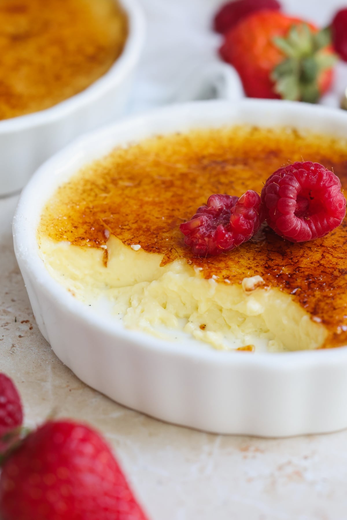 A close-up of crème brûlée in a white ramekin, topped with raspberries. The caramelized sugar crust is cracked, revealing creamy custard beneath—a tempting scene for any creme brulee recipe. Fresh strawberries are scattered around the dish.