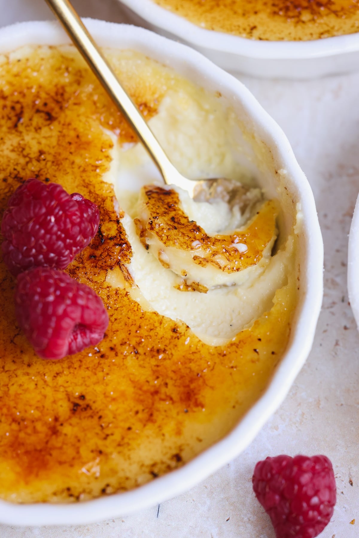 A close-up of crème brûlée in a white ramekin, topped with caramelized sugar and fresh raspberries. A gold spoon has broken through the crispy top, revealing creamy custard.