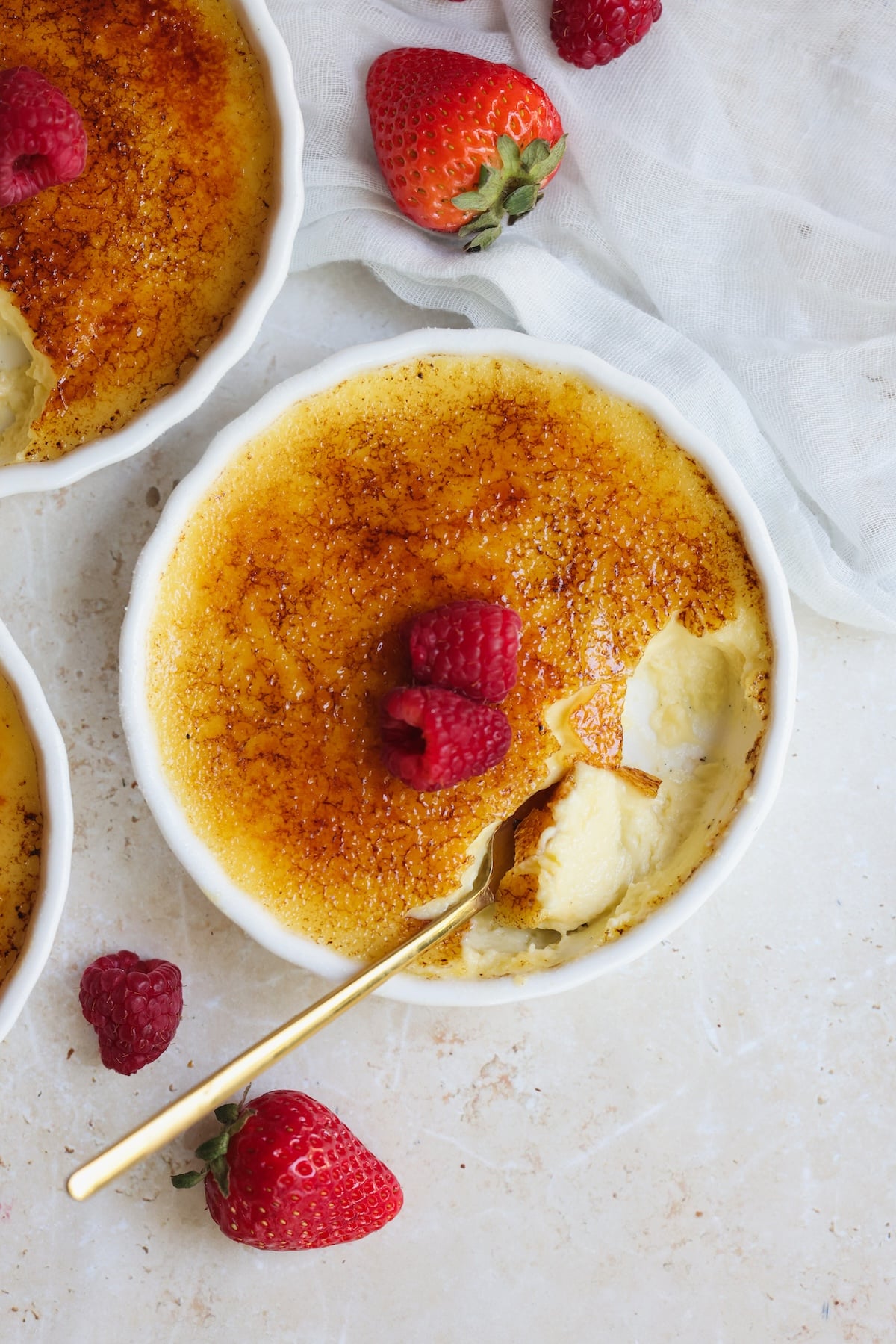 A ramekin of crème brûlée with a caramelized sugar top, partially eaten, garnished with fresh raspberries. A gold spoon rests inside, strawberries and raspberries scattered nearby.