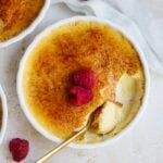 A round dish of crème brûlée, made from a classic Creme Brulee recipe, features a caramelized sugar top and two fresh raspberries. A spoon reveals the creamy custard beneath the crisp layer, with a white cloth partially visible nearby.