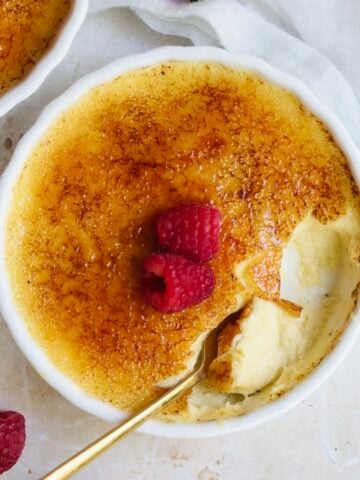 A round dish of crème brûlée, made from a classic Creme Brulee recipe, features a caramelized sugar top and two fresh raspberries. A spoon reveals the creamy custard beneath the crisp layer, with a white cloth partially visible nearby.